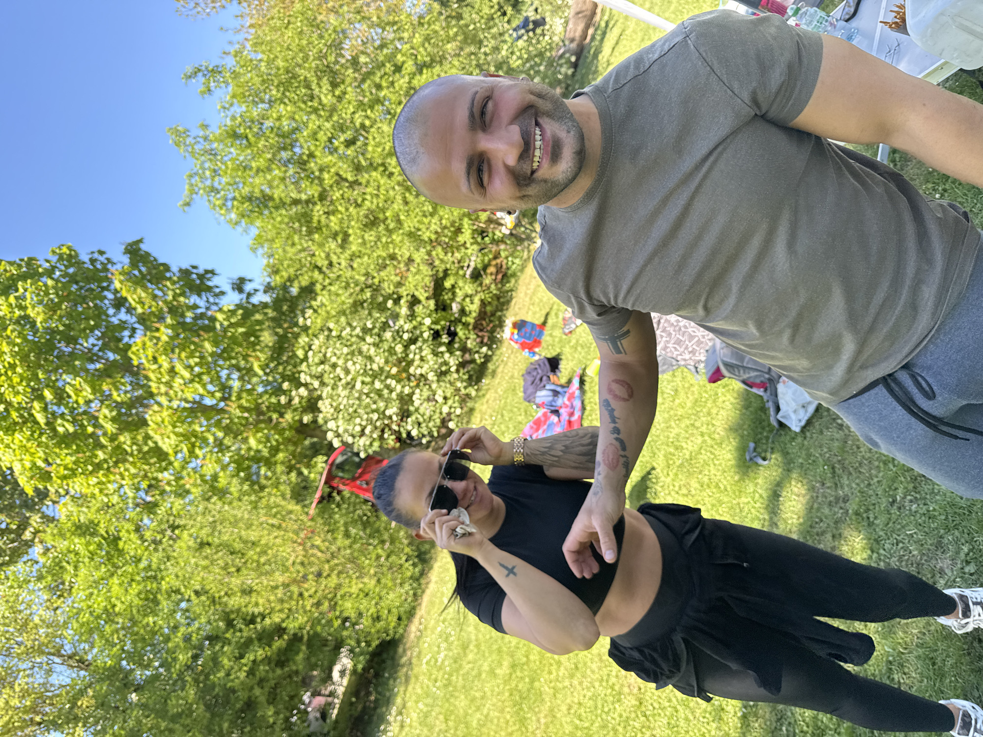 An outdoor, eye-level, tilted shot captures two smiling adults, a man and a woman, in a grassy park or garden setting in Ditzingen, Germany, under a bright blue sky.
The man, positioned in the upper right, has a shaved head, a dark beard, and wears an olive green t-shirt and gray sweatpants. He grins widely, looking directly at the camera. A dark, possibly geometric, tattoo is visible on his left forearm.
The woman stands below him, to the left, wearing a black crop top and black leggings with a ruffled black skirt overlay. Her dark hair is pulled back, and she has sunglasses pushed up onto her head. She holds a small white object in her hand and smiles, looking slightly away from the camera. Her right forearm features distinct red lip-print tattoos, and a small cross tattoo is visible on her left forearm, along with a gold-toned watch.
The background is dominated by vibrant green trees with dense foliage, indicating a sunny day. The ground is covered in green grass. Various personal items, including bags, bottles, and what appears to be a white table and a red stroller or wagon, are scattered in the background, suggesting a casual outdoor gathering or picnic. Both individuals appear happy and relaxed. No visible text is discernible on clothing or objects.