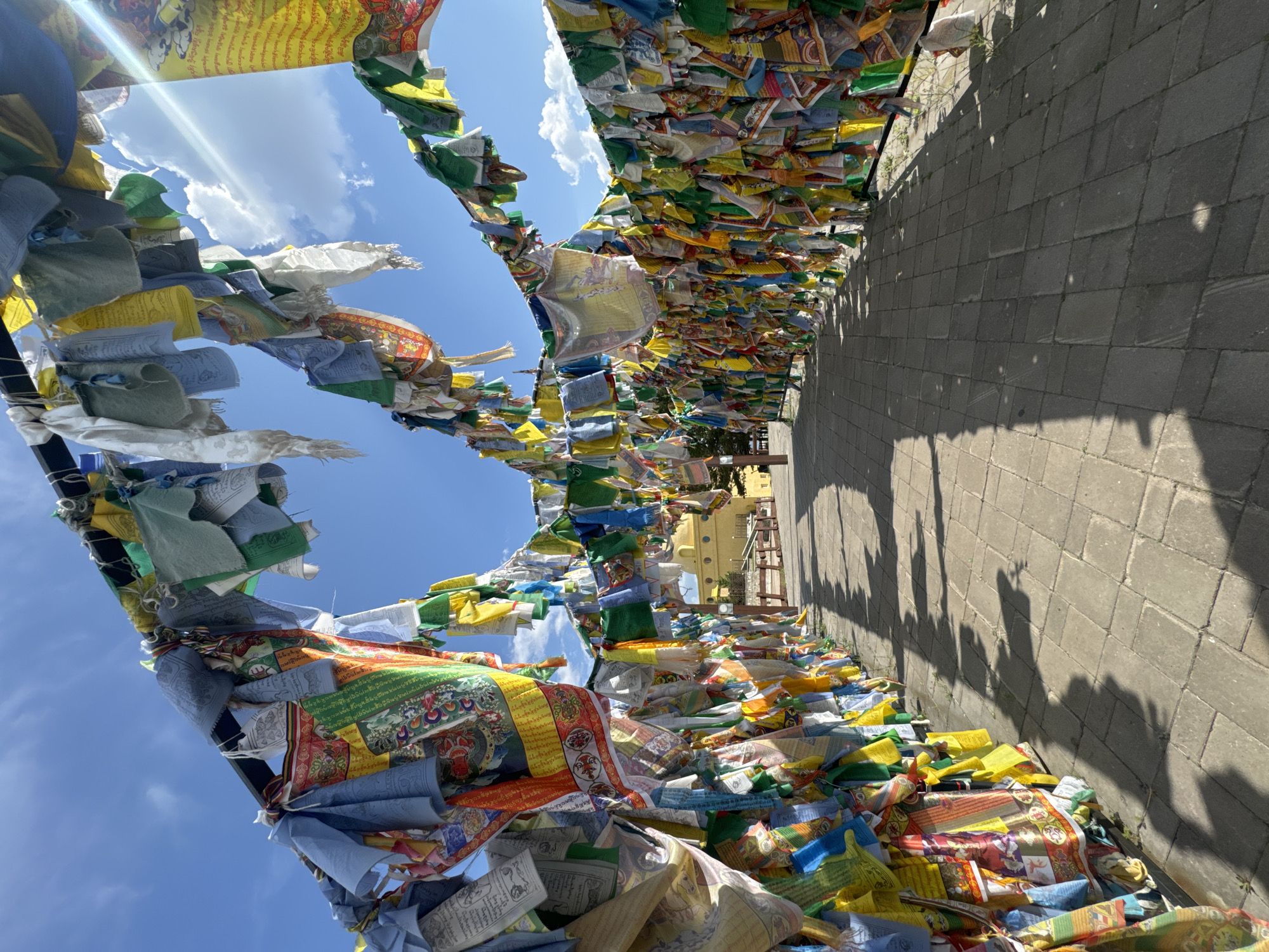 A pathway lined with colorful prayer flags leads towards a yellow building under a blue sky with scattered clouds. The prayer flags, predominantly in blue, white, green, red, yellow, and patterned designs, are strung on metal poles, creating an archway over the path. The pathway is paved with grey interlocking bricks. In the distance, a round, yellow building with a domed roof is visible. Sunlight streams from the right, casting shadows of the flags and poles onto the brick pathway. The scene appears to be outdoors, possibly at a temple or monastery, given the presence of prayer flags. The geographical context provided is the city of Ulan-Ude, Russia, a location known for its Buddhist cultural heritage.