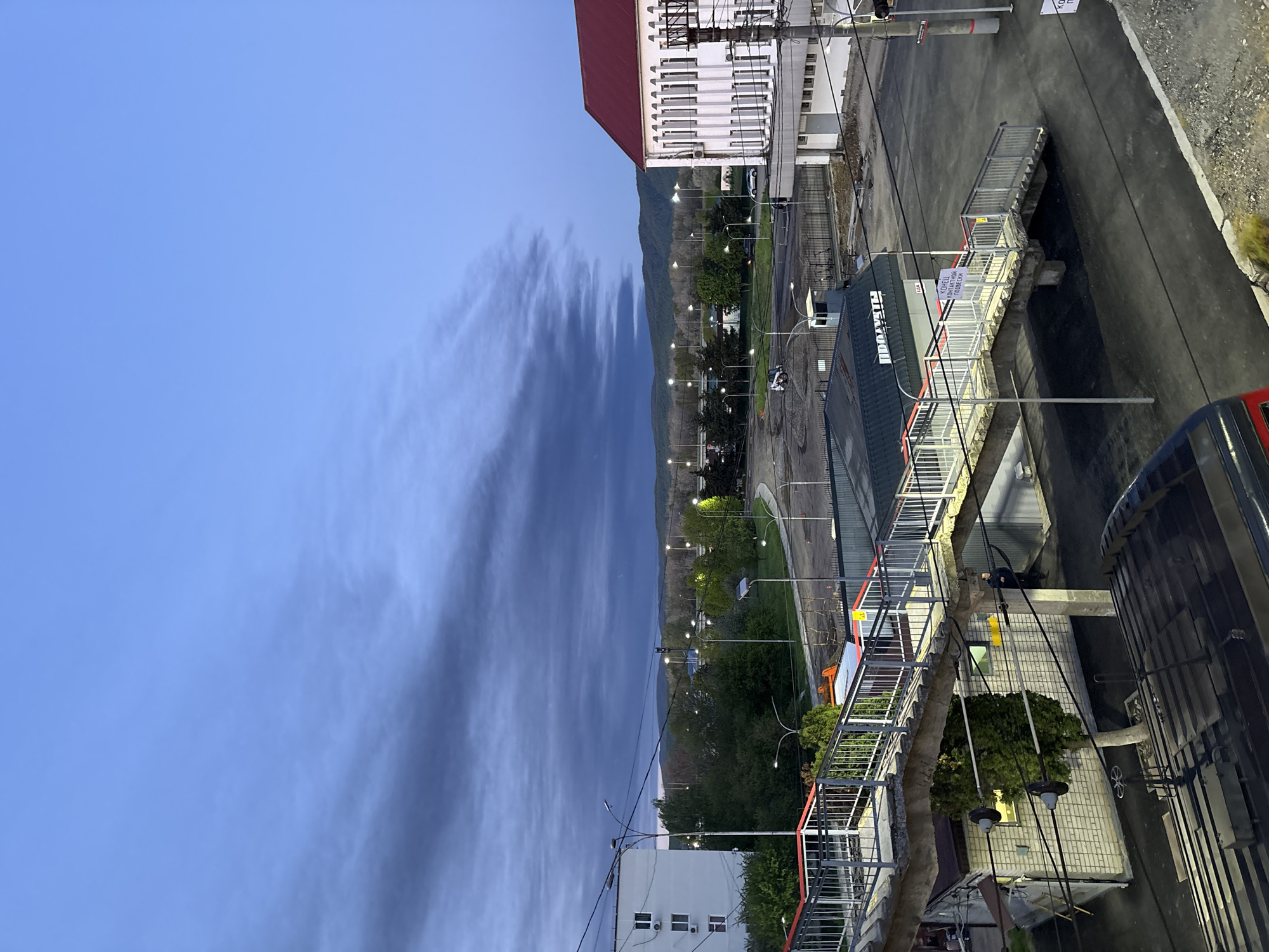 The image, rotated correctly, depicts an urban landscape at dusk or early morning, characterized by a predominantly blue sky with some wispy, elongated clouds. In the distance, dark green mountains rise against the horizon.
In the midground, numerous streetlights are illuminated, casting a glow on roads and green areas with trees and grass. A large, white building with a distinctive red roof is prominent on the left, featuring multiple windows and some visible infrastructure like power lines. To its right, a grey building with a darker roof is visible. Further right, a dark bus or large vehicle is partially seen.
The foreground features various structures and infrastructure elements. A metal stairway with white railings and red accents ascends towards a small building or kiosk. On a white sign with a red border attached to a pole or structure, Russian text is visible: "КОНТЕЙНЕРЫ", "СБОРНИК", "ОТХОДОВ", which translates to "CONTAINERS", "COLLECTION POINT", "WASTE". Another sign on the grey building has the partially visible word "ПОД'ЕЗД" (Entrance). The name "МОРД'АР" (MORDOR) is also visible on the side of this building, suggesting a business or establishment name. Power lines crisscross the scene, attached to poles and buildings. A person in dark clothing is visible near a building or stairway in one of the cropped views. The ground consists of asphalt roads and concrete surfaces.
