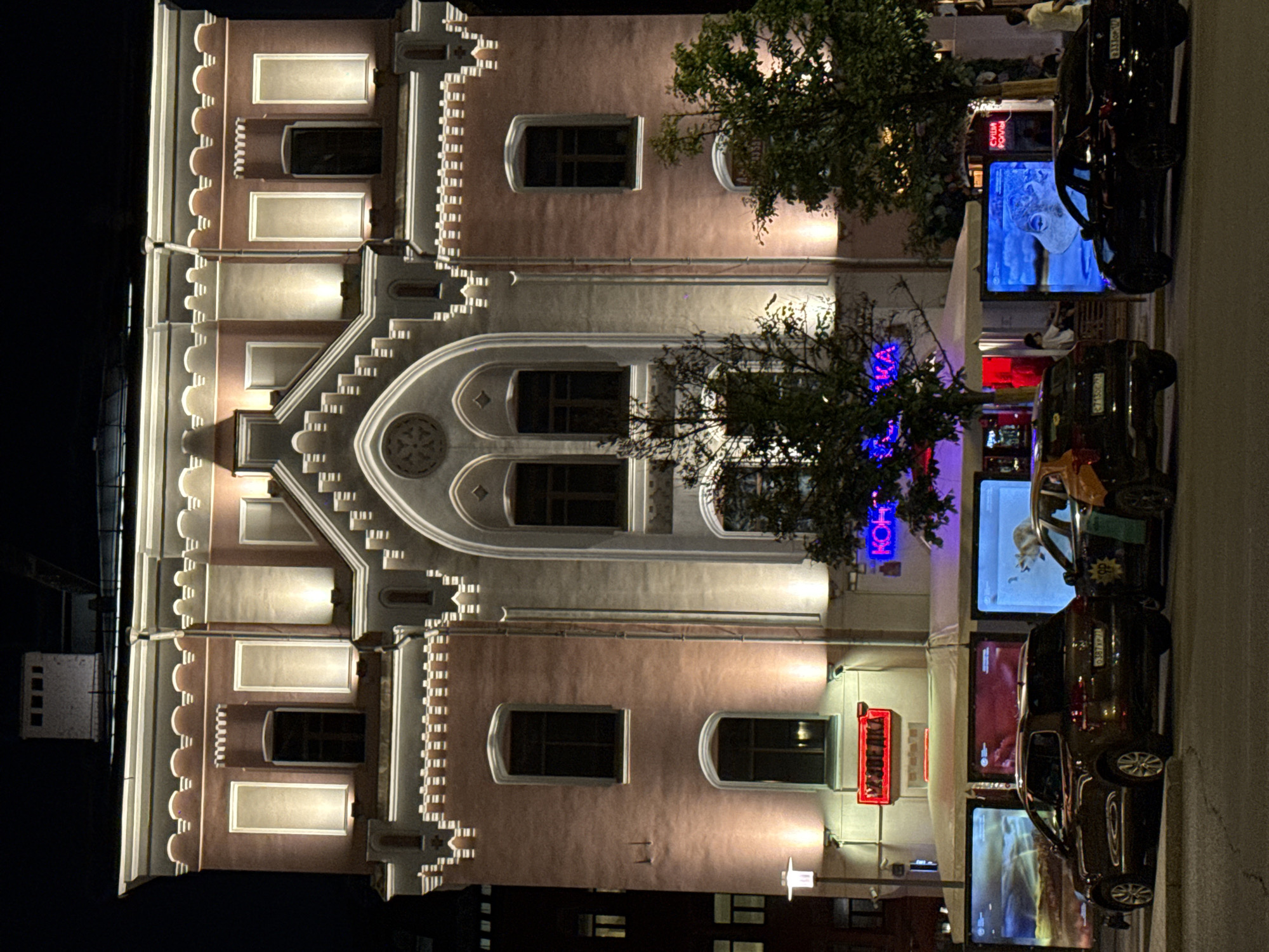 The image depicts a multi-story building at night, likely located in Moscow, Russia based on the Cyrillic text visible on signs. The building has a distinct architectural style with decorative detailing around the windows and along the roofline. It is illuminated by internal lighting shining through the windows and external lights highlighting its facade.
The foreground shows a street scene with several dark-colored cars parked alongside the building. Digital screens are visible along the street, possibly displaying advertisements or information. Trees partially obscure the view of the building in some areas. The street appears to be well-lit with streetlights and light spilling from the building and screens.