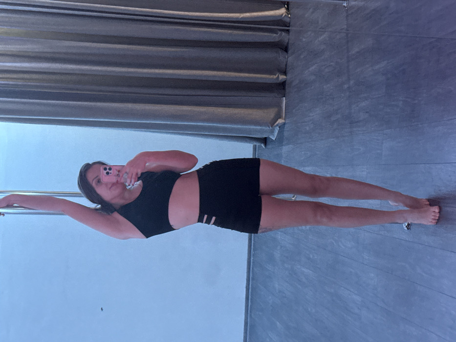 A woman is posing with one arm extended towards a pole, looking into her phone as she snaps a selfie. She is wearing a black sports bra and shorts. The background features curtains along the top edge and a gray floor with a slight sheen. The wall behind her is a neutral, light color.
