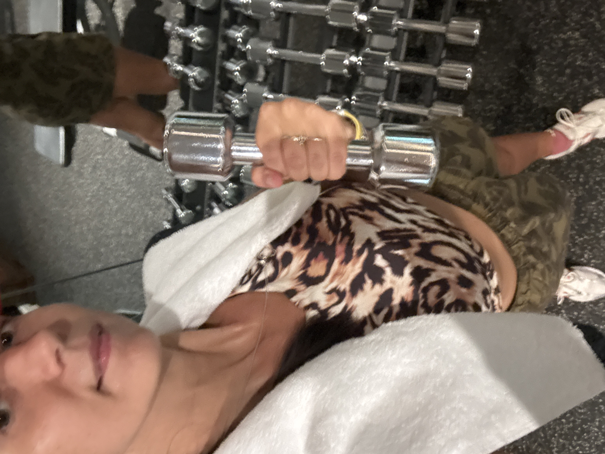 A woman is lying on her back in a gym in Kuta Selatan, Indonesia, holding a dumbbell in her right hand. She is wearing a leopard print sports bra and camouflage shorts. A white towel is draped across her chest and shoulders. A rack of dumbbells is visible behind her. She is wearing white sneakers. The floor is grey with a speckled pattern.