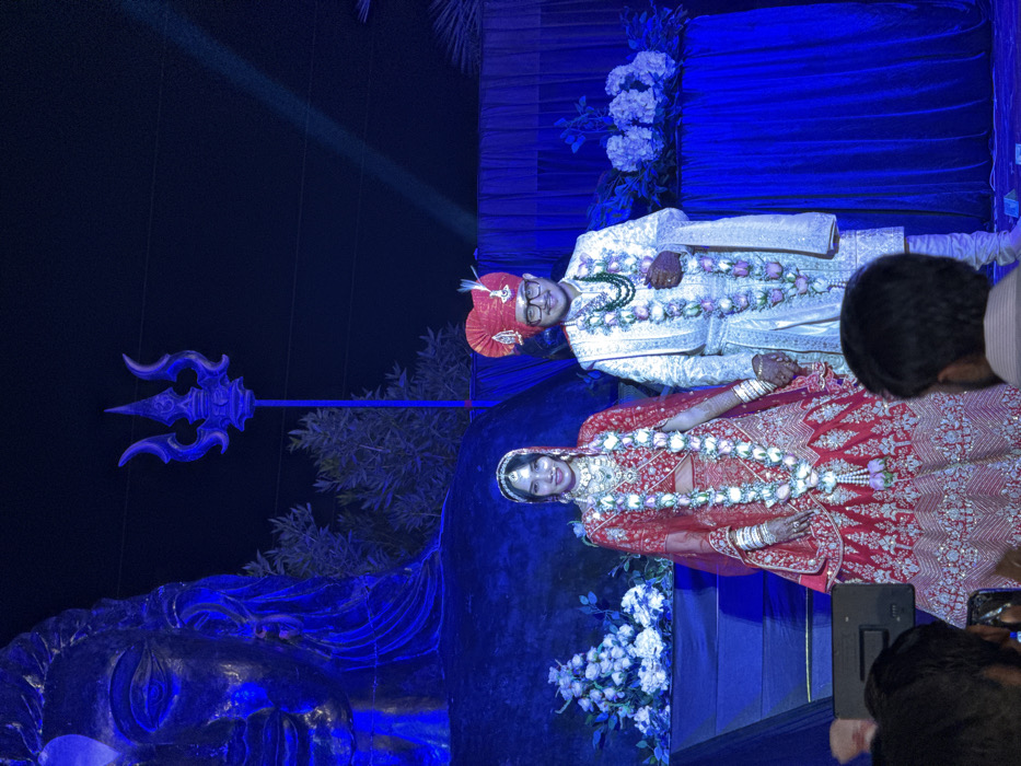 A bride and groom, adorned with garlands, are photographed against a backdrop of blue light, a reclining figure, and a trident. The bride wears a red dress, while the groom is in white and orange. People hold up their phones to capture the scene. White flowers and foliage decorate the background.