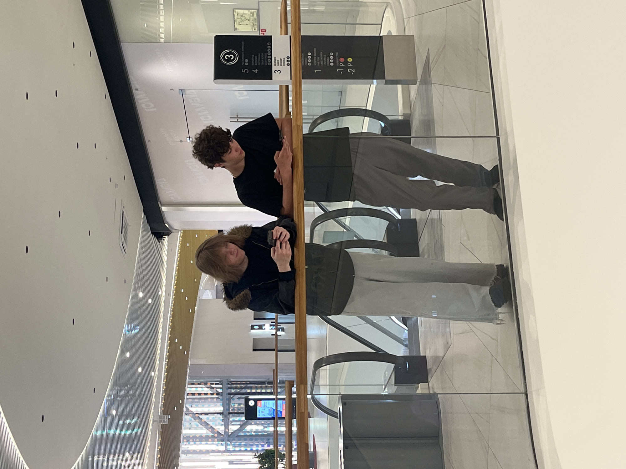 This image depicts two individuals in the Galeria Shopping Mall in Saint Petersburg, Russia.
The scene captures two young males leaning against a clear glass and wooden railing on an upper floor, both engrossed in their smartphones. The person on the left has long, light brown hair and wears a dark jacket with a fur-lined hood, a dark shirt, and light grey wide-legged trousers frayed at the ankles, paired with dark shoes. The person on the right has short, curly dark brown hair, dressed in a black short-sleeved t-shirt, dark grey trousers, and dark shoes. Both individuals are looking down at their phones, displaying no direct interaction with each other or their immediate surroundings beyond leaning on the rail.
To the right of the individuals stands a tall, black and white information sign displaying floor numbers (3, 5, -1, -2) and a list of establishments in Cyrillic, including "ЗОЛОТОЕ ЯБЛОКО" (Golden Apple), "М. ВИДЕО" (M. Video), and "РЕСТОРАНЫ С ТЕРРАСОЙ" (Restaurants with Terrace). Behind the right individual, a curved glass wall reflects the words "ГОЛЛИВУД" (Hollywood) and "ПРЕМЬЕРА МЕЧТЫ" (Dream Premiere). In the background, dark escalators descend, and a bright, modern interior with multiple levels is visible. The ceiling features diverse lighting elements, including recessed spotlights, strip lighting behind horizontal metallic slats, and a section with individual small lights over a gold-toned surface. The floor is light-colored tile. A grey cylindrical object, possibly a waste bin, is partially visible to the left. The overall environment is brightly lit, spacious, and modern, indicative of a large commercial complex.
