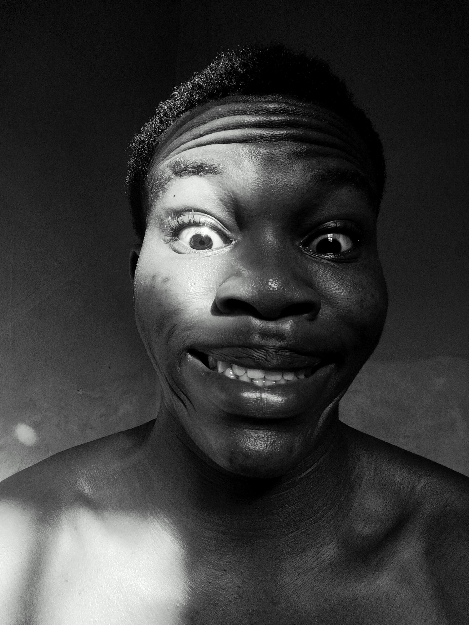 A black and white, close-up portrait depicts a person with dark skin. The individual has a wide, toothy grin and bulging eyes, creating a somewhat exaggerated and comedic expression. The forehead is furrowed, and there is noticeable shine on the skin, likely due to lighting. The person is bare-chested, with part of the shoulders visible at the bottom of the frame. The background is a muted gray, with some areas appearing darker than others, suggesting an indoor setting with uneven lighting. Location: Unknown, Unknown.