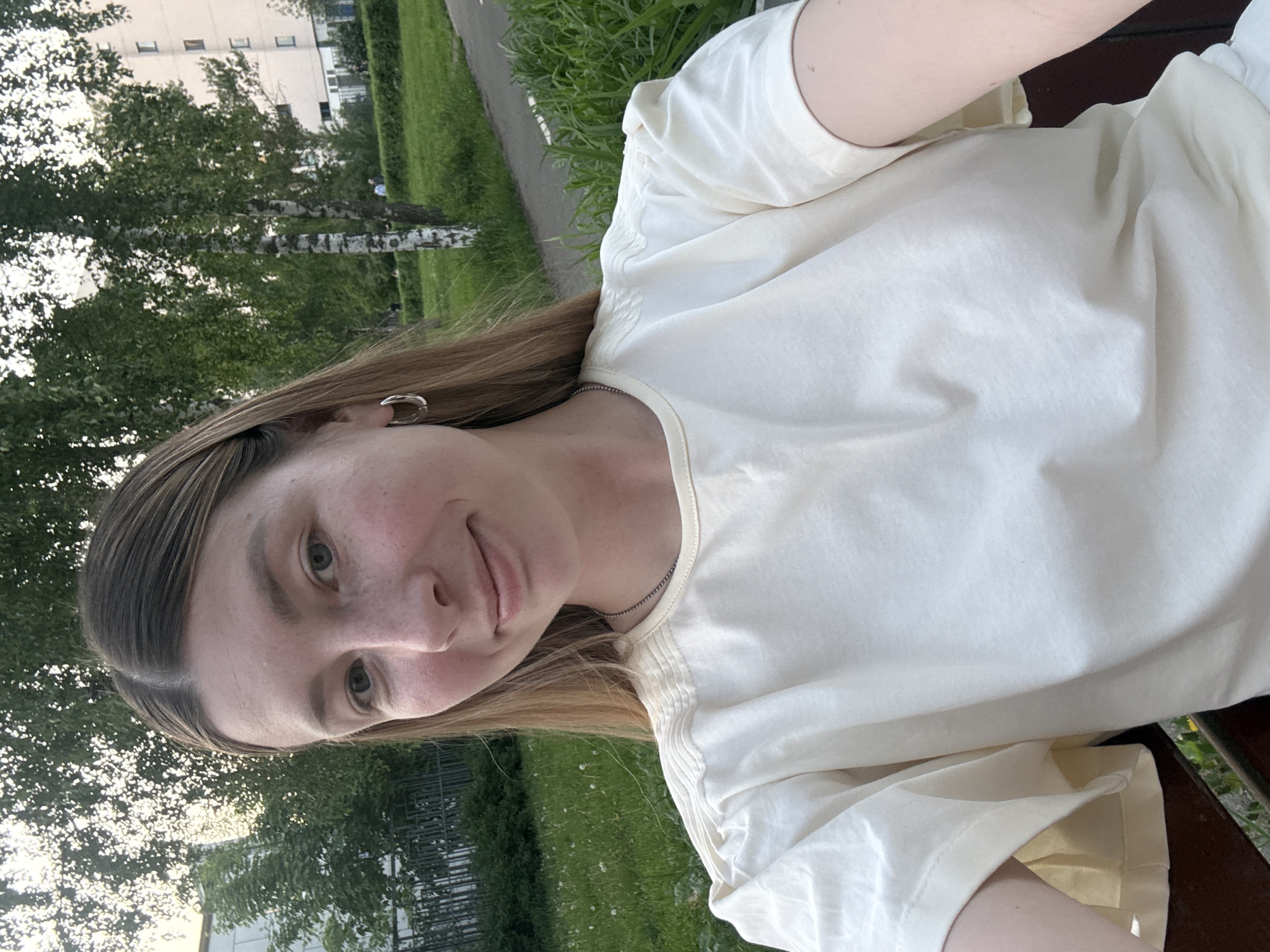 A young, fair-skinned woman with light brown hair is looking directly at the camera. She is wearing a casual, light-colored t-shirt. A simple hoop earring is visible in her left ear. The background shows a green, grassy area with trees and what appears to be a multi-story building in the distance. Given the architecture and general appearance, the location is potentially somewhere in Moscow, Russia, but there is no irrefutable evidence to definitively confirm this.