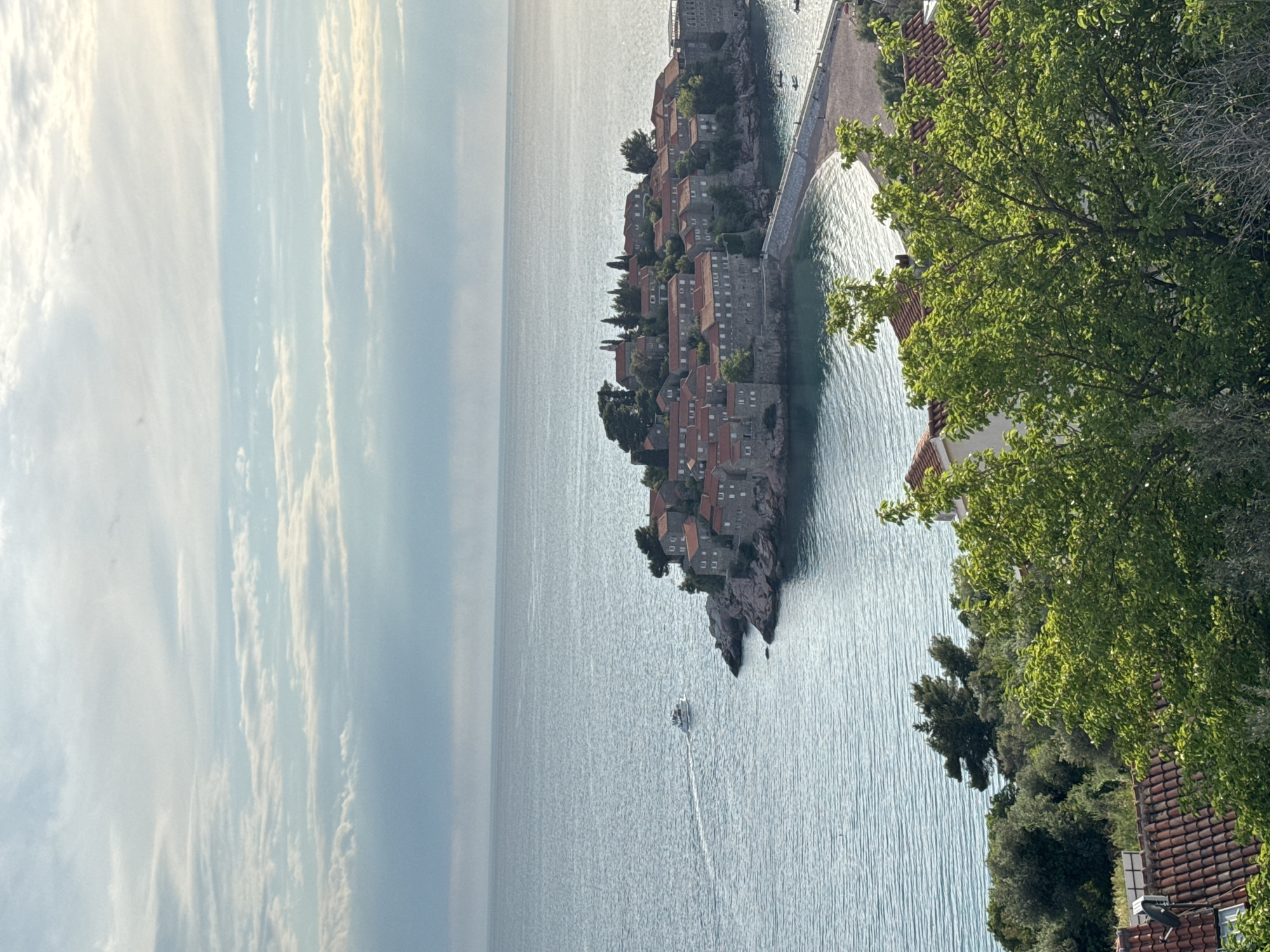 A daytime, elevated, angled shot captures Sveti Stefan, Montenegro. The blue-gray sea stretches to the left, meeting a sky with striated clouds. An island with densely packed stone buildings with red tile roofs is connected to the mainland by a causeway. A boat moves in the water near the island. Green trees are on the right, with rooftops visible beneath.