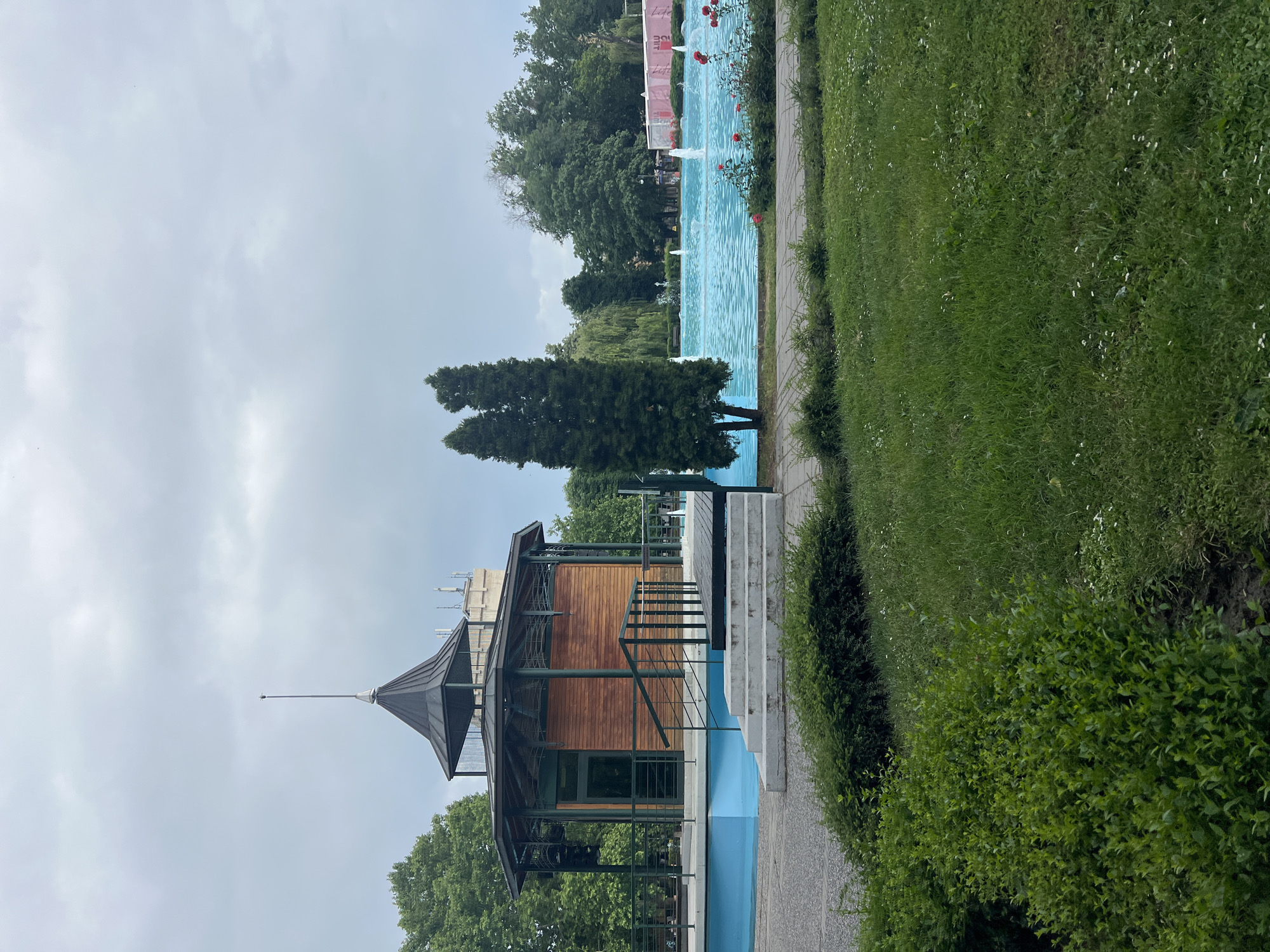 This image captures a serene park landscape, featuring a long, light blue artificial water channel or fountain that stretches across the middle ground. Several small water jets create white sprays along the length of the channel.
On the left side of the water feature stands a distinctive pavilion-like building. It boasts wooden paneling on its exterior walls and is topped with a dark, multi-faceted metal roof that culminates in a slender, pointed spire. Steps lead up to a platform adjacent to this structure.
To the right of the water channel, a well-defined path runs alongside an expansive area of vibrant green grass, interspersed with small white flowers. Manicured green bushes form a border along the path. A few red roses add splashes of color near the water's edge on the right.
The background is filled with a variety of trees, including two tall, slender evergreen trees positioned prominently near the center-left, and a denser cluster of deciduous trees further back. The sky above is overcast and a pale grey, indicating a cloudy daytime setting. No individuals are visible, contributing to the tranquil atmosphere of the scene. In the distant background, a sign or banner partially displays the word "Life" in red lettering. This picturesque setting, with its distinctive fountain and architecture, is located in Plovdiv, Bulgaria, aligning with features found in parks such as the Tsar Simeon Garden.