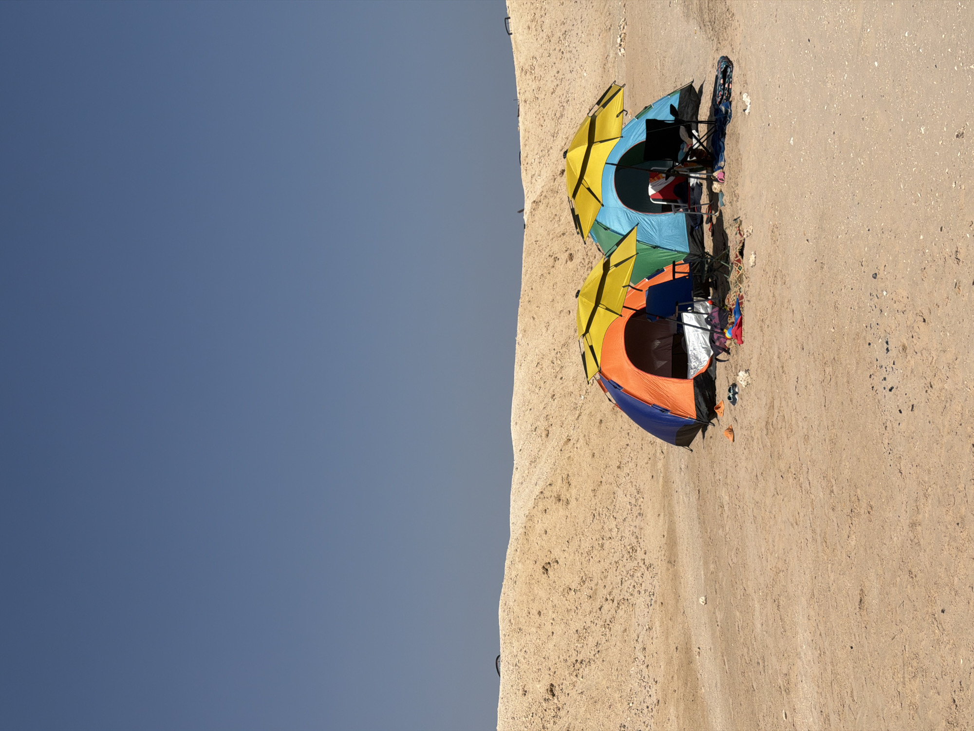 This image depicts a sandy, arid landscape under a clear blue sky, likely in Sharm El Sheikh, Egypt. Two colorful tents, one orange and blue, the other green and blue, are set up on the sand. Each tent is partially covered by a yellow umbrella. In front of the tents, beach chairs are visible, along with some beach accessories like towels and bags. The ground is sandy with scattered small rocks and debris. In the background, a large sand dune rises, and on top of it, there appear to be distant vehicles or structures. The scene suggests a recreational setting, possibly a beach or desert campsite.