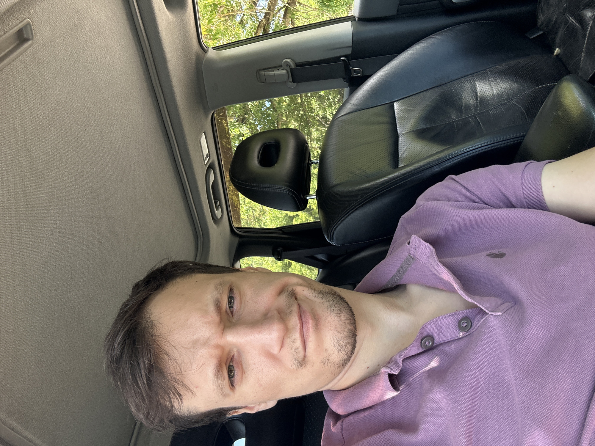 A man with short, dark hair and light green eyes is captured in a close-up selfie taken inside a car. He is wearing a light purple polo shirt. His facial expression is somewhat skeptical, with a slight smirk and furrowed brow. The interior of the car is visible, including a black leather headrest and seat, a grey headliner, and a portion of the dashboard and door panel. Sunlight streams in, illuminating the scene and casting shadows. The background through the car window shows lush green foliage, suggesting an outdoor, possibly rural or suburban, setting. There is no indication of the specific location in the city of Makiv, Ukraine.