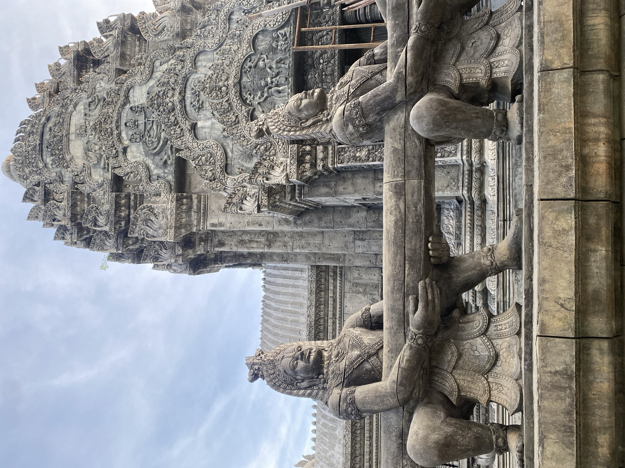 The image displays a section of a highly ornate stone temple or architectural structure, likely located in Ban Haad Rin, Thailand, against a lightly cloudy sky. In the foreground, two large, carved stone figures, resembling guardians or mythical beings, are prominent. Each figure is adorned with an elaborate crown, jewelry, and detailed clothing, all meticulously sculpted from stone. They are depicted holding onto a thick, horizontal stone beam or staff, integrated into the architecture. The figure in the lower part of the image (when oriented correctly) looks slightly upwards with a serene yet powerful expression, showcasing intricate details like patterned scales or feathers on its lower body.
Behind these guardian figures, the building's facade is richly decorated with elaborate stone relief carvings. These feature numerous smaller figures, possibly deities or mythological scenes, interwoven with complex floral motifs and geometric patterns that extend across multiple tiers of the structure. The weathered grey stone suggests considerable age and historical significance. The scene is illuminated by the soft, diffused light of a cloudy sky, which highlights the intricate textures and depths of the ancient craftsmanship. No visible text is present. The overall impression is one of profound cultural heritage and architectural grandeur.