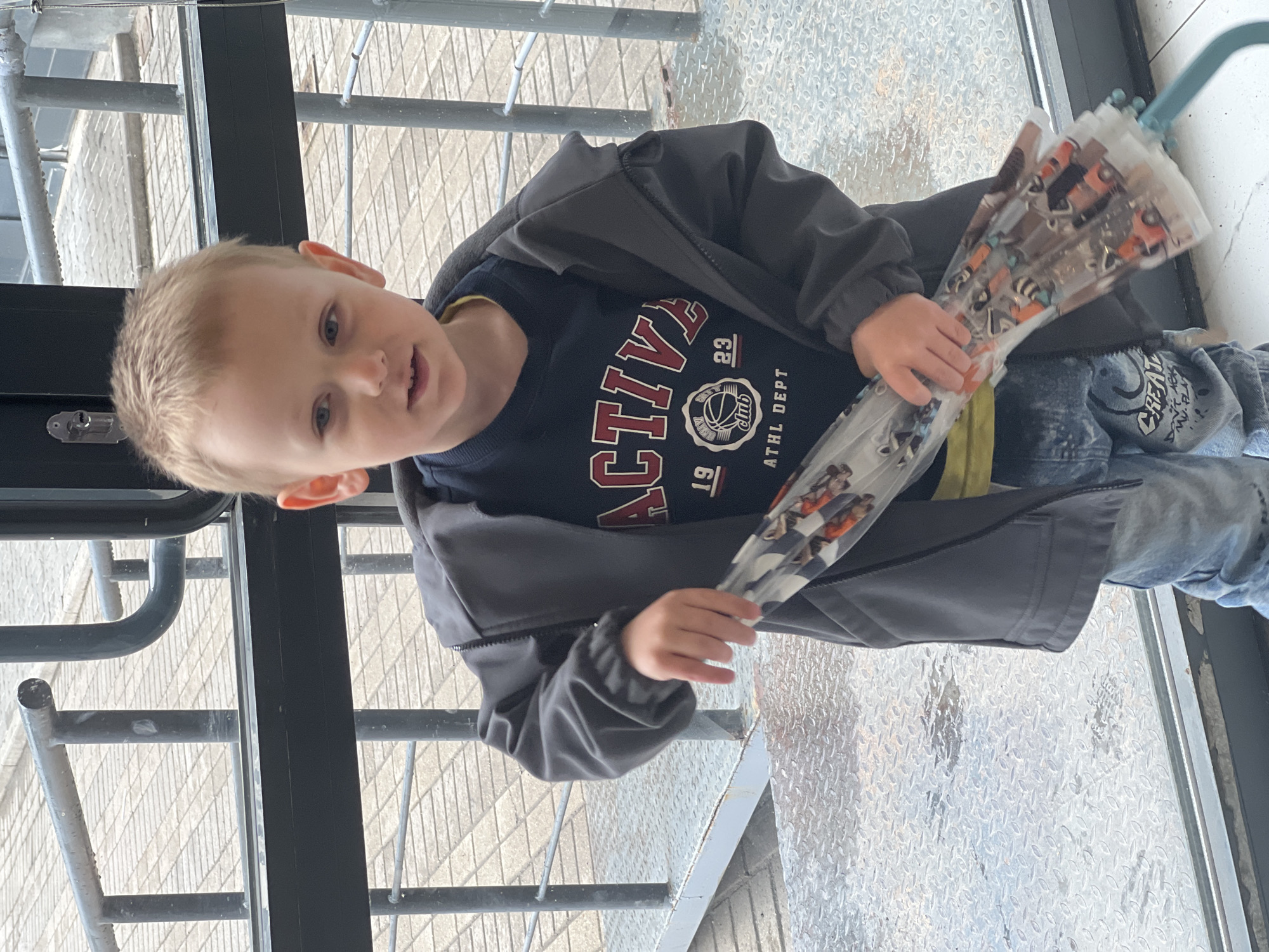 A young boy stands indoors on a metal grate floor, likely within a building or enclosed structure given the interior setting. He is dressed in a dark blue sweatshirt featuring a graphic design of "Active" with a basketball theme, a gray jacket, and blue jeans. The boy holds a transparent umbrella with cartoon character prints. He appears to be facing the camera, with a slight smile or open mouth. The background includes metal structural elements, potentially a staircase or platform. Based on the location in Mytishchi, Russia, as requested, the surrounding architecture might include industrial buildings or public spaces commonly found in urban Russian settings.