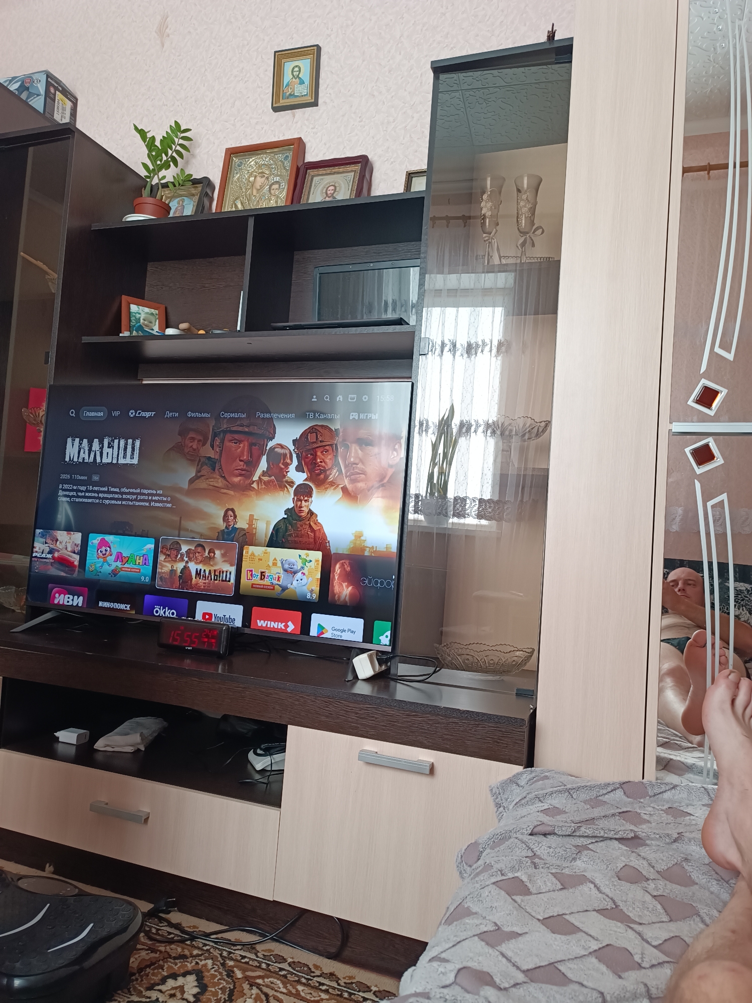 In Vuhlehirsk, Ukraine, a television displays a movie poster with the title "МАЛЫШ". The screen also shows a selection of other movies and applications. To the left of the television, a tall, dark brown cabinet holds several framed religious icons and a potted plant. To the right of the television, a glass-fronted cabinet displays two decorative vases. The scene is set in a living room with a patterned rug on the floor. In the reflection of the glass cabinet, a person's legs and feet are visible, lying on a blanket. A digital clock on the shelf below the television reads 15:55.