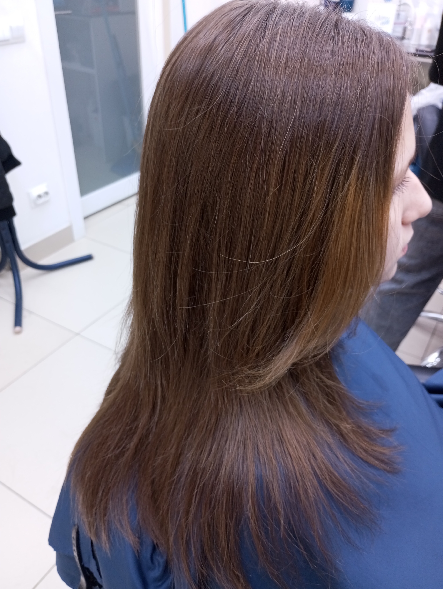 The image shows a woman with long, straight, light brown hair in what appears to be a hair salon. She is wearing a blue cape or smock, likely to protect her clothing during a hair appointment. The location appears to be inside a building with white tile flooring and white walls. There is a white door and what looks like salon equipment in the background. The scene is brightly lit, suggesting indoor lighting. There's no information to place it in Izhevsk, Russia.