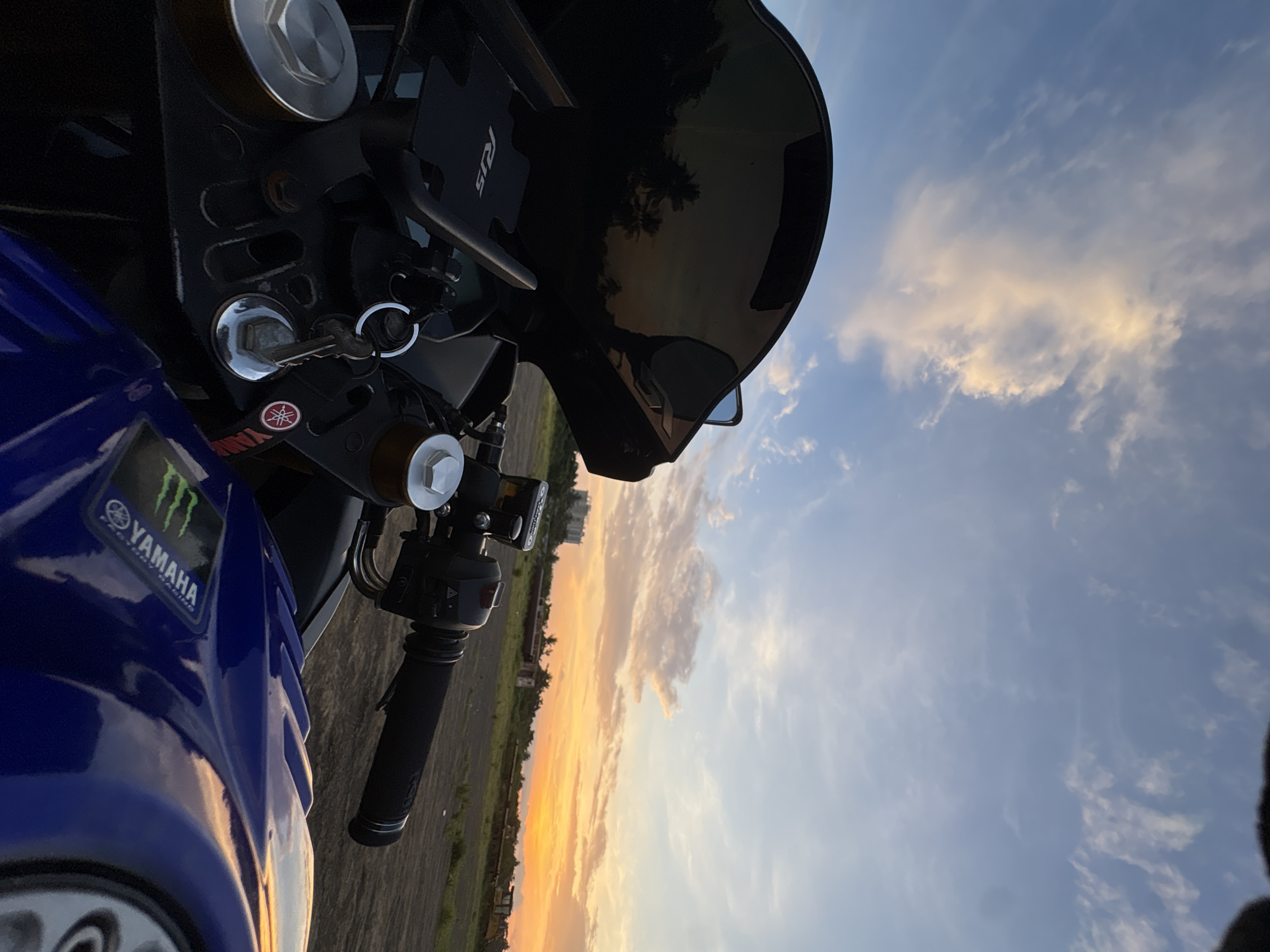 A close-up, low-angle shot captures a motorcycle parked outdoors, likely in an open area on the outskirts of Davao City, Philippines, given the tropical vegetation visible in the reflection and the distant industrial elements. The foreground is dominated by the motorcycle's blue fuel tank, adorned with a "Yamaha Factory Racing" decal, and its handlebars with a textured grip. The central portion of the image shows the motorcycle's instrument panel, featuring a black windscreen with reflections of palm trees and a colorful sunset sky. A set of keys hangs from the ignition. The background reveals a dusky, flat landscape with scattered derelict structures and construction equipment, under a sky filled with dramatic clouds painted in hues of orange, yellow, and blue, indicative of sunset. The lighting suggests it is late afternoon or early evening. There are no people or animals visible in the scene.