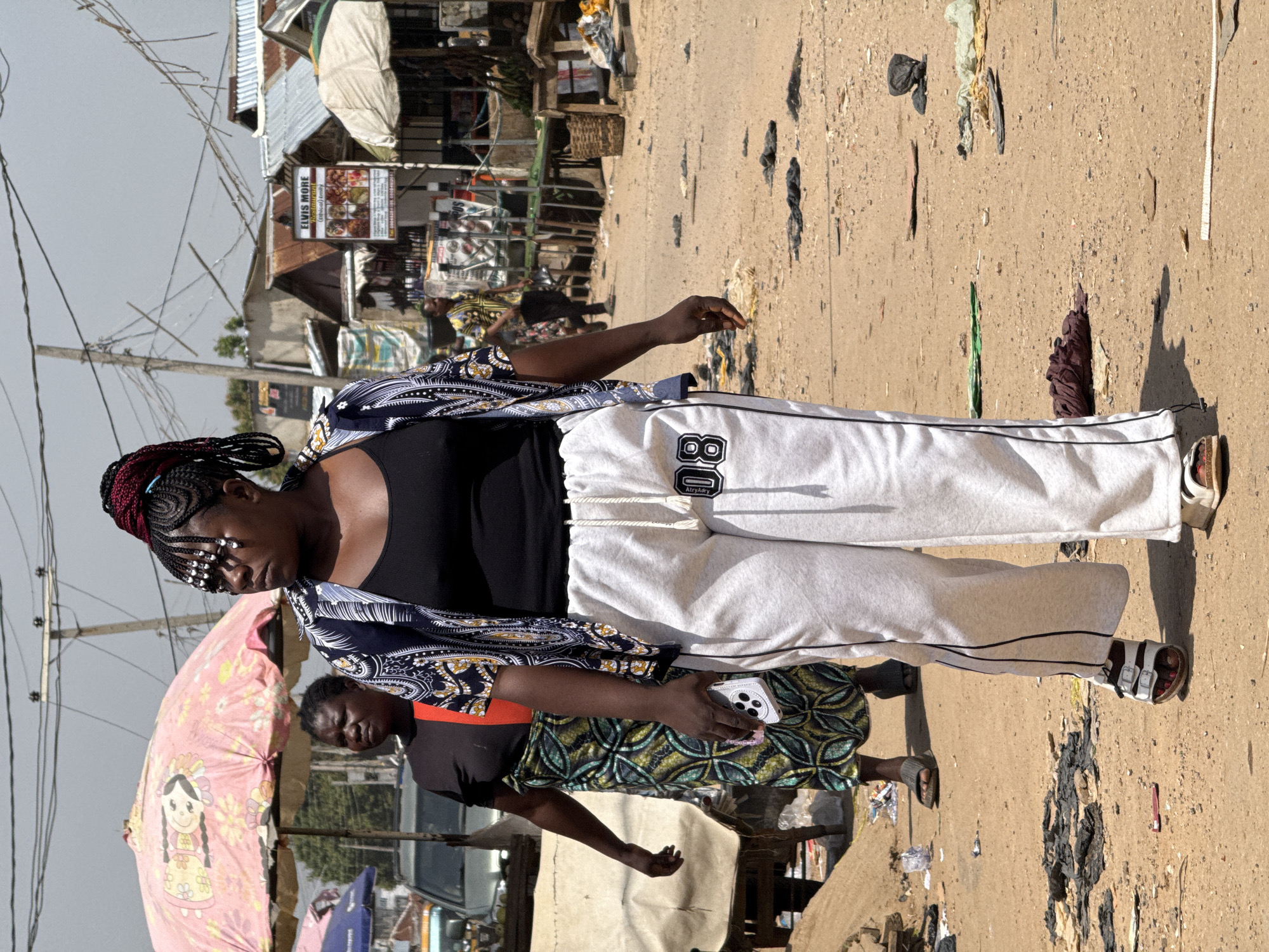 This media file depicts an outdoor scene in what appears to be a market or busy street in Abuja, Nigeria. The primary subjects are two women, with one prominently featured in the foreground. She is wearing a black tank top, a patterned blue and white kimono-style jacket, and white sweatpants with "80" printed on the leg. She has braided hair with some red accents and decorative beads around her forehead. Her gaze is directed downwards, and she is holding a white smartphone.
Behind her, another woman is visible, wearing a dark top and a patterned green and yellow dress. She also appears to be looking down. A pink umbrella with a cartoon character design is partially visible on the left side of the frame, suggesting it might be a vendor's umbrella or personal item.
The setting is a dusty, unpaved street with various stalls and buildings in the background. There are piles of trash and debris scattered on the ground, indicating a potentially unkempt environment. The architecture visible in the background includes corrugated metal roofs and simple structures, characteristic of some urban areas in Nigeria. Overhead, a complex network of wires suggests an active but perhaps disorganized electrical infrastructure.
The lighting and shadows indicate it is daytime, likely mid-morning or afternoon, with bright sunlight casting distinct shadows. The overall atmosphere appears to be one of everyday activity in a bustling, possibly informal, marketplace.
Visible text includes a sign that reads "ELVIS MORE" with "family" written below it, and what appears to be a menu or advertisement. Another tag on the sweatpants reads "AtryAdry". The context of Abuja, Nigeria, helps ground the scene in a specific geographical and cultural environment.