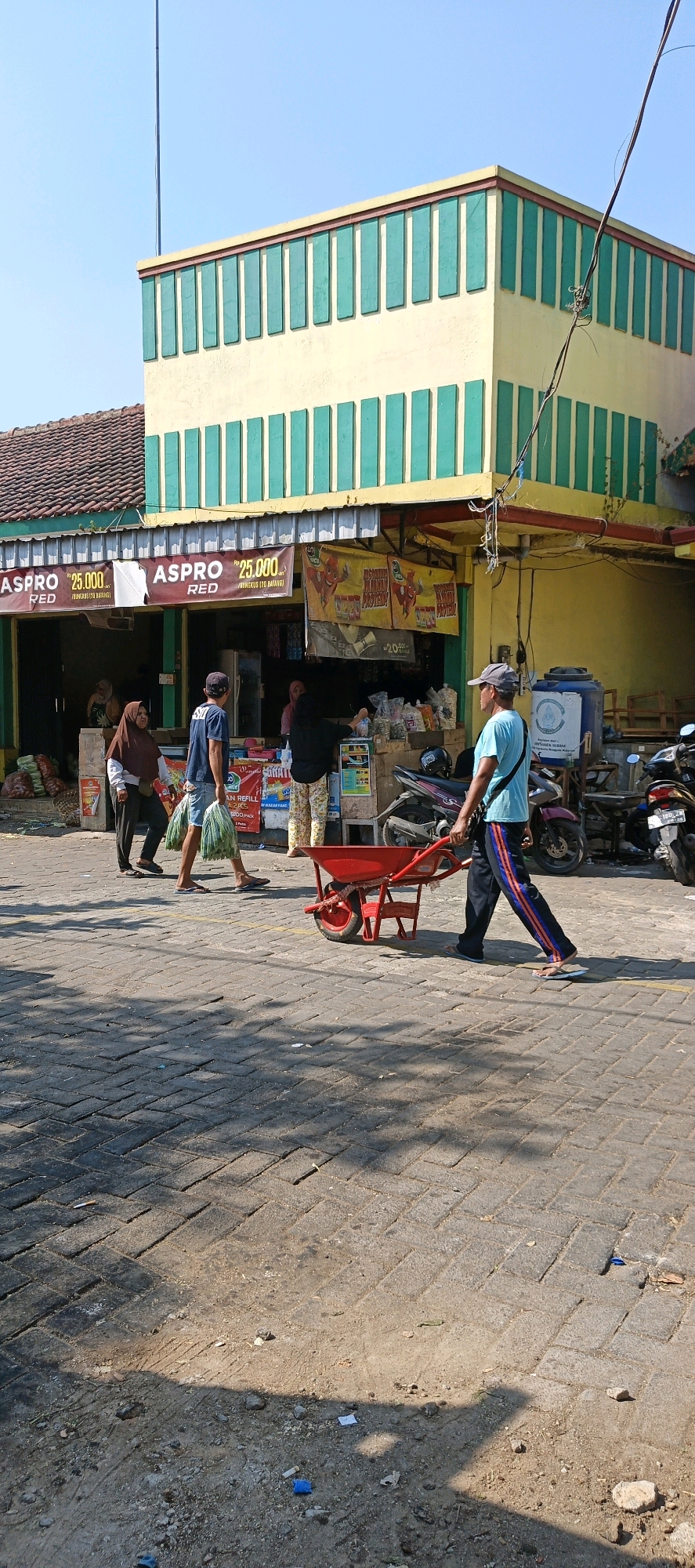 In the city of Klaten, Indonesia, on a sunlit day, a man is pushing a red wheelbarrow along a brick-paved road in front of a yellow and green building that appears to be a shop. The building has a tiled roof and signs advertising "ASPRO RED" with prices. Several people are visible near the shop entrance, some carrying bags. A woman in a brown hijab is walking away from the shop, and another woman in a black headscarf is interacting with someone behind a counter. A man in a blue t-shirt and denim shorts is walking towards the shop, carrying two large green plastic bags filled with produce. Motorcycles are parked to the right of the shop. The ground is dusty with scattered debris. The overall scene depicts a typical day at a local marketplace.