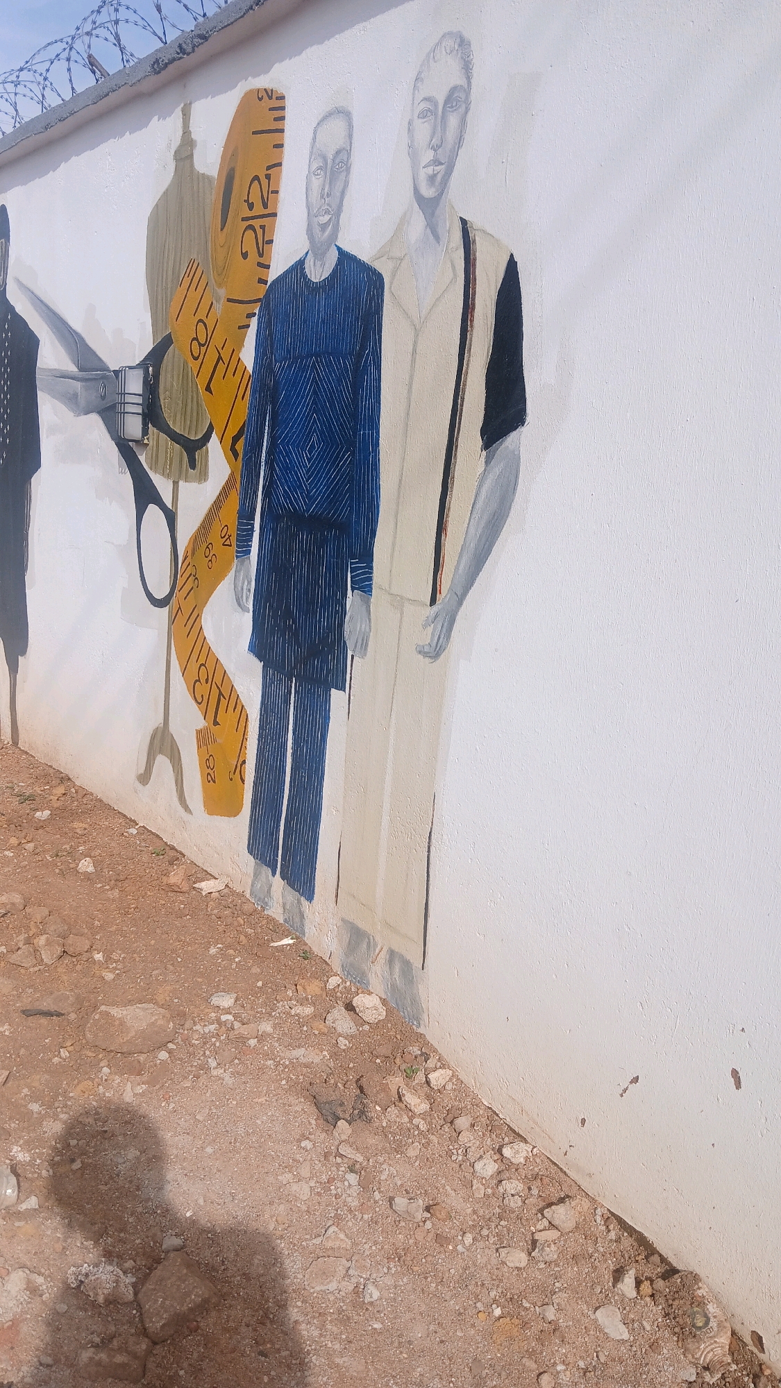 The image shows a mural painted on a white wall in Gura Top, Nigeria. The mural depicts two figures dressed in stylized clothing, possibly representing tailors or fashion designers. To the left of the figures are elements of a tailor's workshop, including a dress form, a large pair of scissors, and a yellow measuring tape with markings. The ground in front of the wall is covered with reddish-brown dirt and small rocks. A shadow of a person is visible on the ground, indicating the presence of a photographer or observer. Barbed wire is visible along the top of the wall, suggesting it is part of a perimeter or security fence. The overall scene appears to be an outdoor setting, likely in a public or semi-public area within the town.