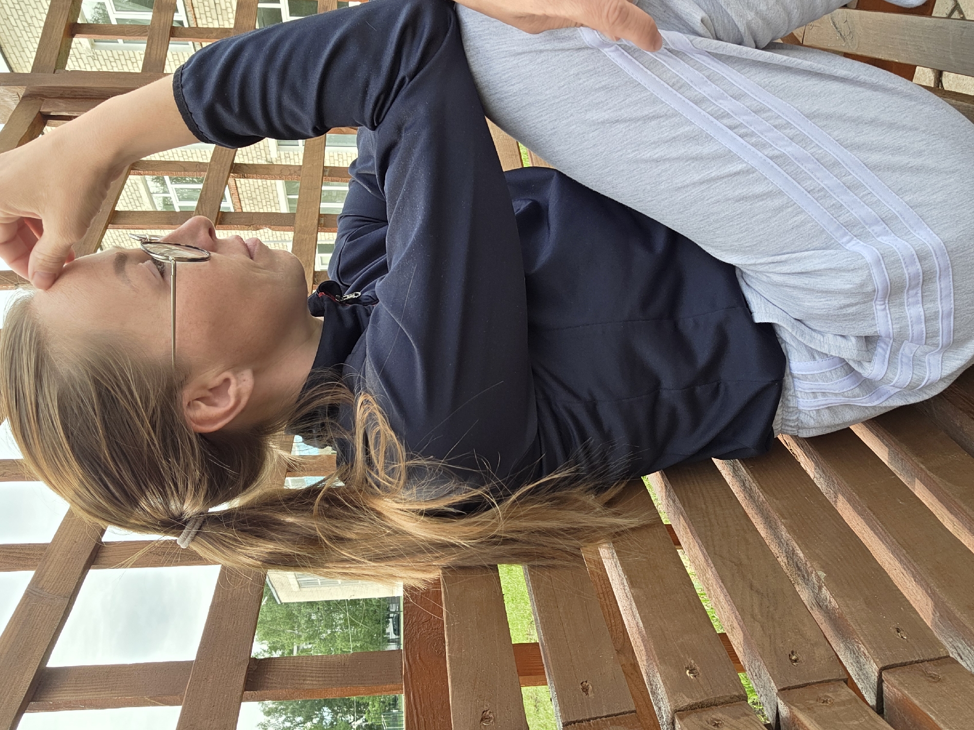 The image shows a woman lying on a wooden bench, possibly in a pergola or gazebo. She is wearing glasses, a dark blue jacket, and gray sweatpants with three white stripes down the side. Her hair is blonde and pulled back into a ponytail. Her left hand is touching her forehead. The scene appears to be outdoors, with trees and buildings visible in the background. The location seems to be in a residential area, possibly in the city of Unknown, Unknown.