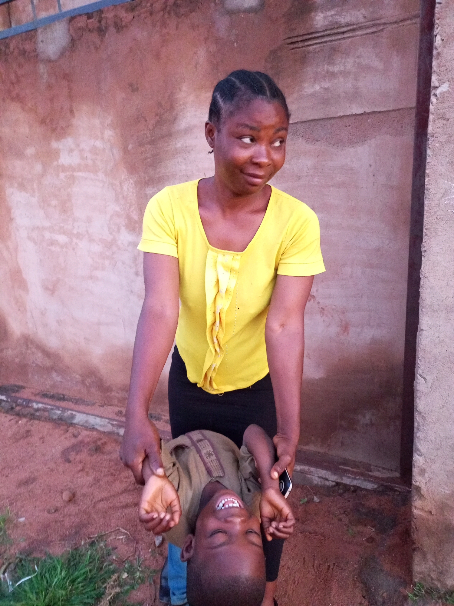 A young Nigerian woman with braided hair and a yellow shirt is holding a young boy upside down by his arms. The boy is laughing and his eyes are closed. The scene appears to be outdoors, with a textured wall in the background and reddish-brown dirt on the ground. There is some grass to the left of the frame. The woman is wearing black pants. The overall impression is one of playful interaction.