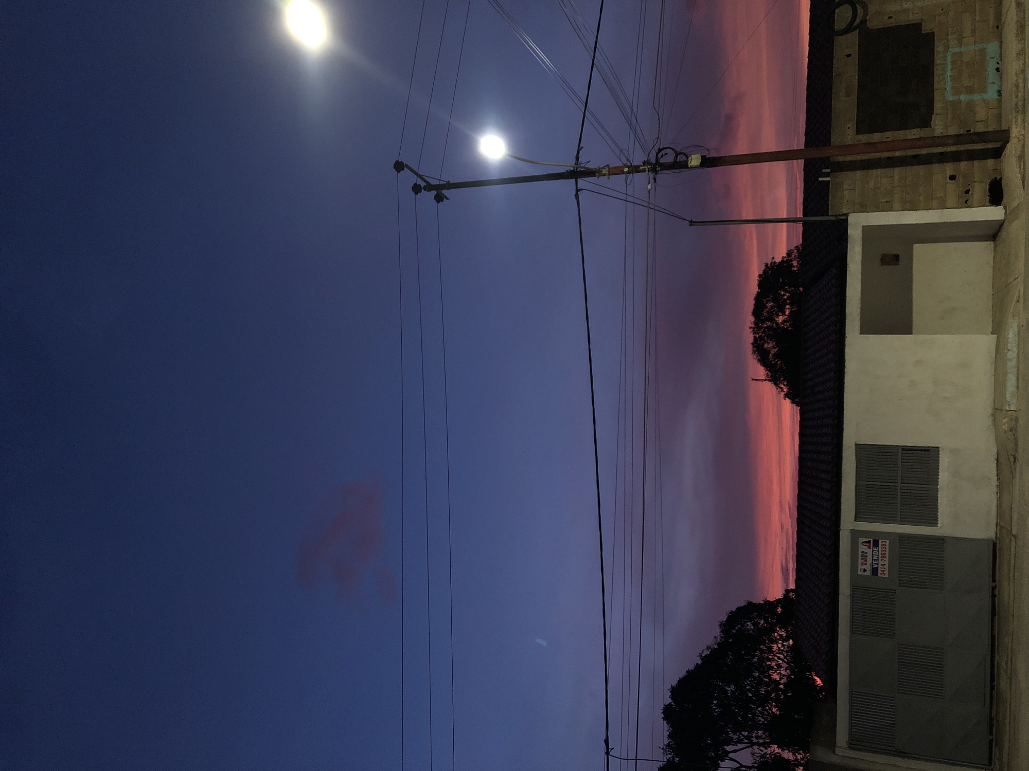 The image depicts a twilight scene in a residential area, likely in Altagracia, Venezuela. The sky transitions from a deep blue to a vibrant pink-orange near the horizon, suggesting either sunset or sunrise. Utility poles with wires crisscross the sky, leading to a street lamp that is illuminated, casting a bright light. On the right side of the frame is a building with a gray garage door featuring a real estate sign. A portion of the building's facade is visible, including a small covered porch. Dark foliage, likely from trees, borders the building's roofline. The overall impression is of a quiet, suburban evening.