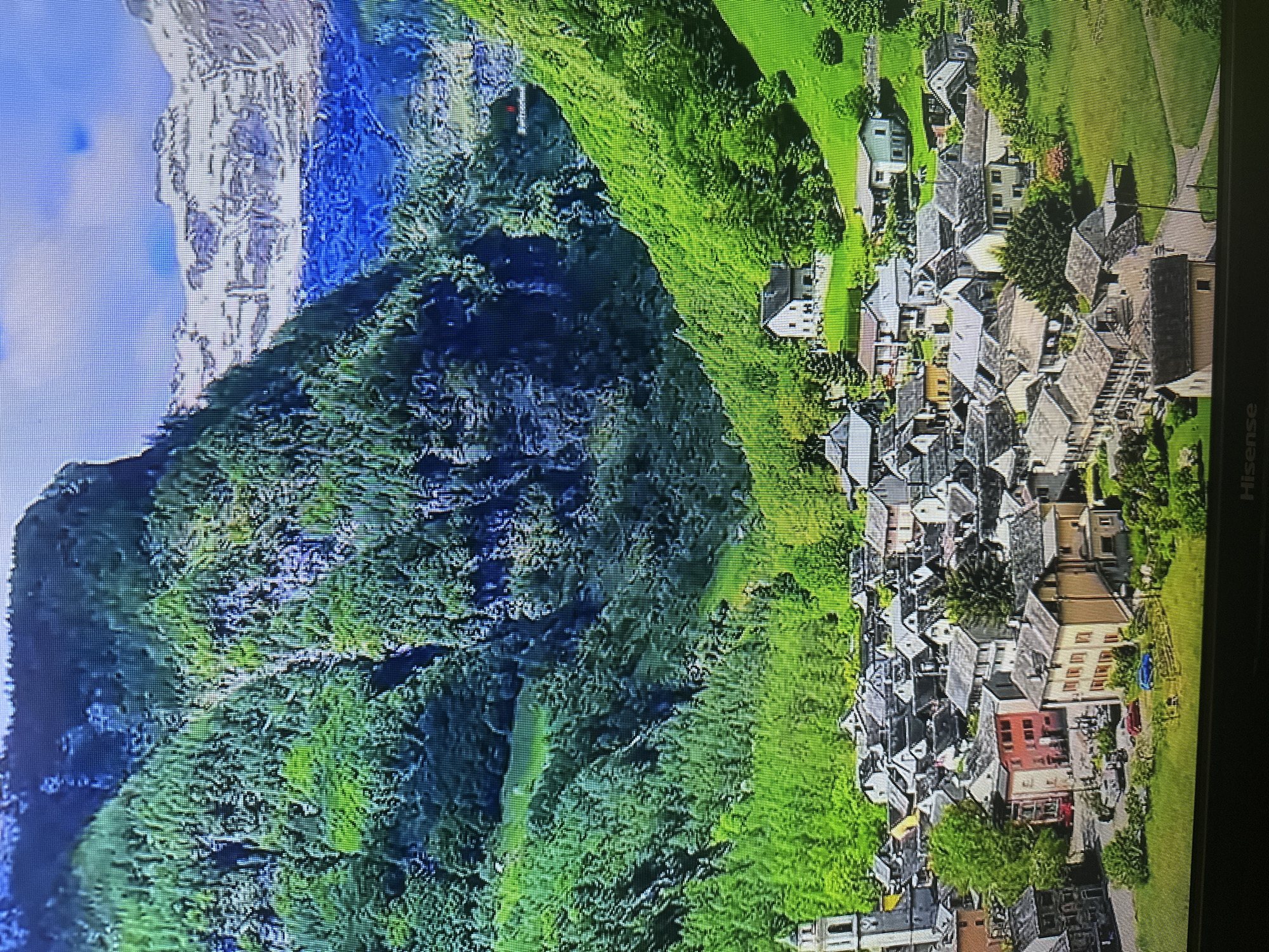 The image shows a scenic mountain village with traditional stone houses. Steep, tree-covered slopes rise behind the village, and in the distance, snow-capped mountains are visible under a blue sky with scattered clouds. A tall bell tower stands out in the village. The scene depicts a peaceful, idyllic setting, characteristic of a rural European mountainous region. The provided context stating this is in Kyiv, Ukraine, is inaccurate given the visual evidence of a mountainous landscape with snow-capped peaks and traditional alpine architecture.