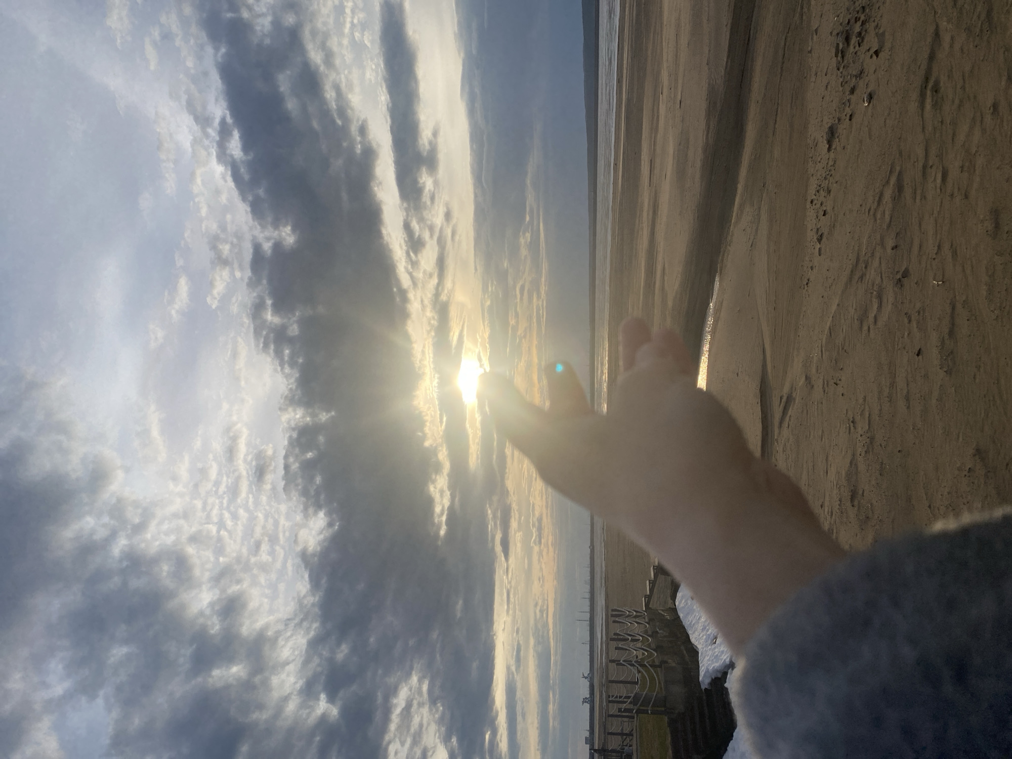 The image captures a beach scene in what appears to be Chaikovsky, Russia, given the context. The sky dominates the view, filled with a mix of cloudy patches and clear sections, with the sun breaking through. A hand is outstretched towards the sun. The shoreline is sandy, and in the background, there's a glimpse of what might be a retaining wall or structure along the beach. There are no other people visible in the frame besides the hand and arm. The environment appears to be lit by the sun.