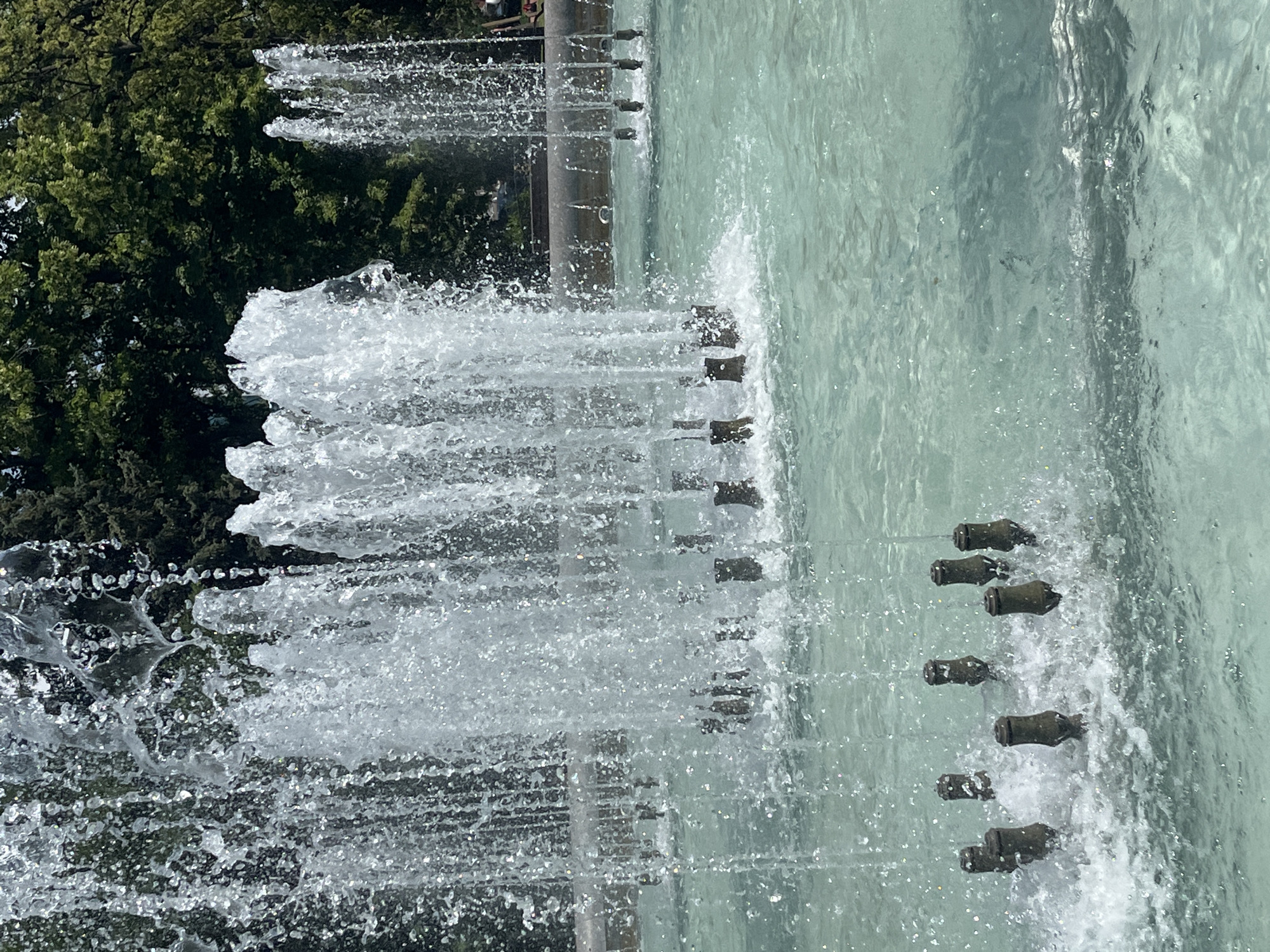 The image depicts a close-up view of a fountain in a park-like setting, likely in Krasnodar, Russia, given the vegetation. Several streams of water are being ejected from submerged nozzles, creating a dynamic display of splashing and cascading water. The water is clear and a pale turquoise color, with sunlight reflecting off its surface, creating sparkling highlights. In the foreground, a cluster of dark, metallic nozzles are visible just above the water's surface, surrounded by turbulent water. Further back, more nozzles are emitting taller, more vigorous jets of water. The background is filled with lush, green trees, suggesting a warm, sunny day. The overall impression is one of a refreshing and lively public space.
