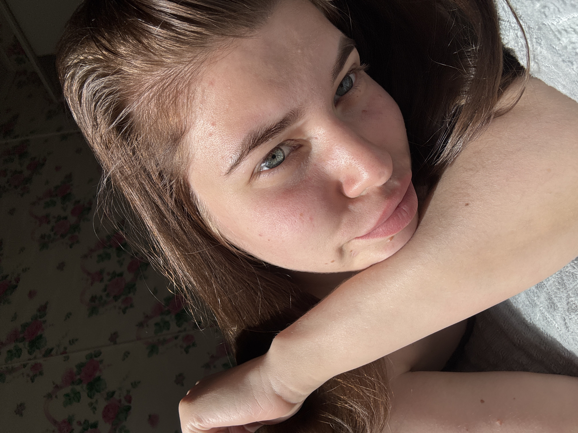 A close-up shot features a light-skinned woman with brown hair and light-colored eyes. Her head is slightly tilted. Her right arm is bent and positioned beneath her head. The background includes a wall with a floral pattern and a portion of a grey fabric. The photo does not depict any elements that indicate the specific location of Мещёры, Russia.