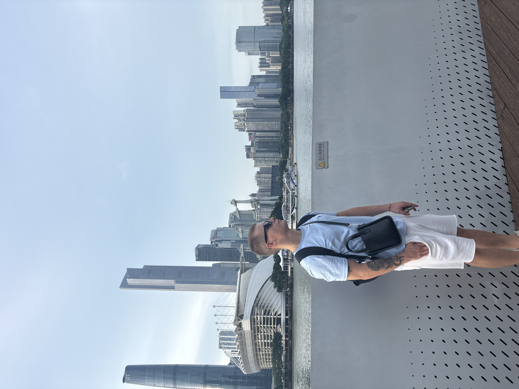 A young man stands on a modern walkway, looking up at a cityscape. He is wearing a light blue t-shirt, white shorts, and sunglasses, with a small black crossbody bag slung over his shoulder. His left arm is adorned with a tattoo. The walkway has a perforated metal railing and a wooden deck on the right side.
In the background, a wide river separates the walkway from a dense urban skyline dominated by numerous tall buildings, including the Canton Tower and the Guangzhou Opera House, indicating the location is likely Guangzhou, China. The sky is clear and bright, suggesting daytime. The overall impression is one of leisure and urban exploration. A warning sign is visible on the railing in Chinese and English, reading "Warning: hot surface."