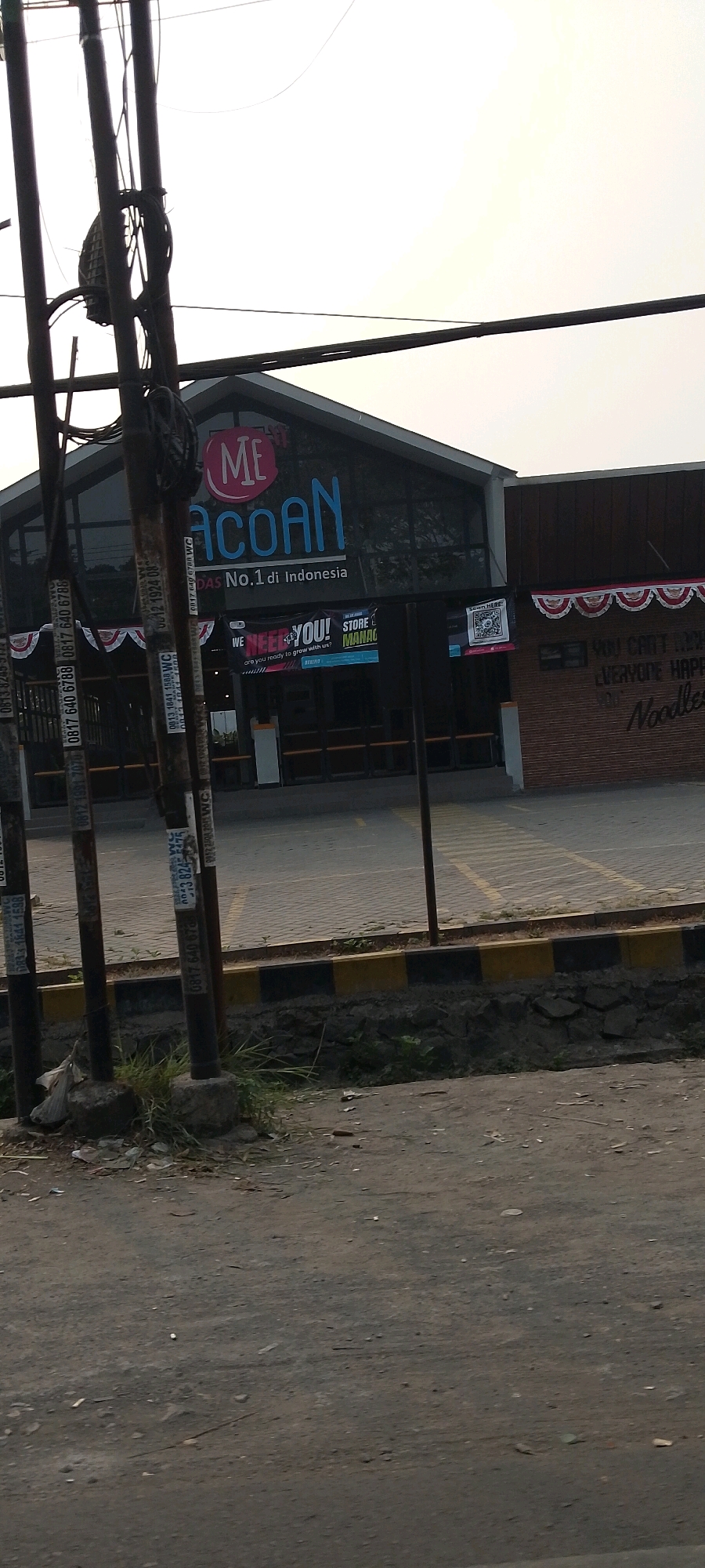 The image shows the exterior of a "MECOAN" store, a fast-food restaurant chain that claims to be "No.1 di Indonesia" (No. 1 in Indonesia). The store is located in the city of Bekasi, Indonesia. The building has a modern design with large glass windows and a dark-colored facade. Above the entrance, the "MECOAN" logo is prominently displayed. A banner hangs in front of the store that reads "WE NEED YOU! STORE MANAGER".
In the foreground, there are several utility poles with electrical wires and some posters attached to them. The ground in front of the store is paved with interlocking bricks, with a yellow and black striped border indicating a safety zone. Beyond the paved area, there is a dirt road with some scattered debris. The sky is overcast, suggesting a cloudy day. The overall scene depicts a commercial area in Bekasi.