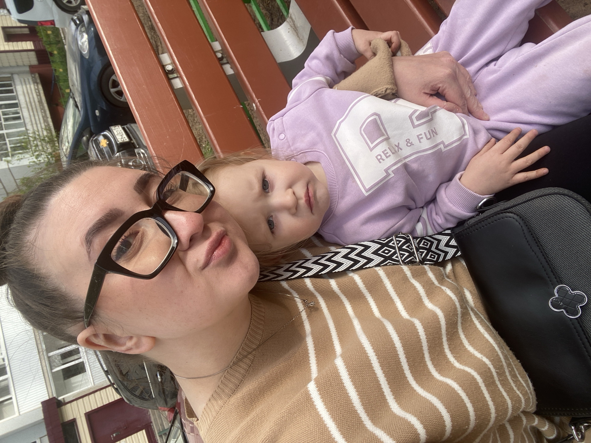 A woman wearing glasses and a tan and white striped sweater is taking a selfie with a young child lying on her lap. The child is wearing a light purple sweatshirt with the words "Relix & Fun" on it. An adult's hand is seen holding the child. A black purse with a clover-shaped decoration sits on the woman's right side. The background shows a brown and green bench, a parked car, and a building. The setting appears to be outdoors. The location is in the city of Chaikovsky, Russia.