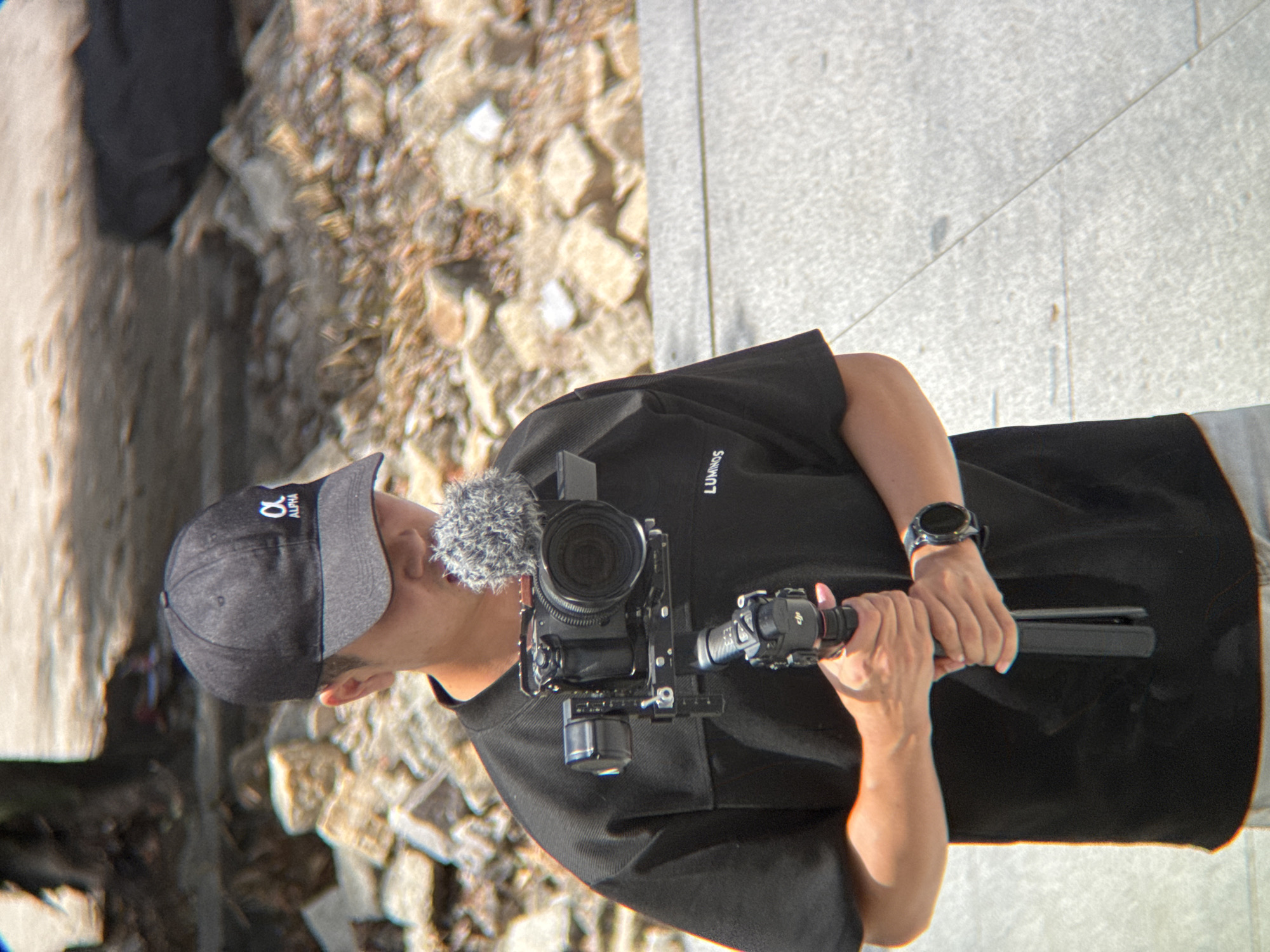 The image shows a person wearing a black baseball cap with an "alpha" logo and a black t-shirt with "LUMINOUS" written on it. The person is holding a camera rig with a microphone attached to it. The rig is mounted on a gimbal. The person is standing on a paved surface, possibly a sidewalk or plaza, with a background of rubble and some foliage. The lighting suggests it is daytime. The provided information indicates the location is Nha Trang, Vietnam, though this is not visually evident from the image itself.