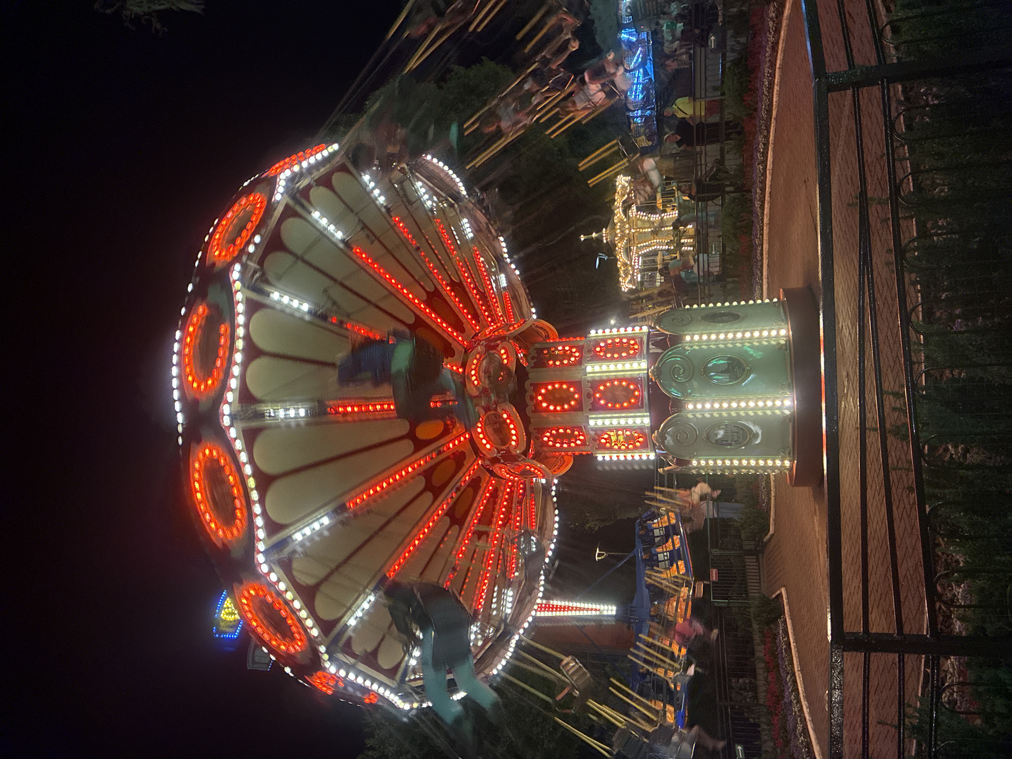 The image depicts a nighttime scene at an amusement park, possibly located in Sochi, Russia, given the context. The central focus is a brightly lit swing ride, characterized by its circular canopy adorned with numerous red and white lights. Chairs with people are seen in motion, blurred by the ride's rotation. In the background, other amusement park rides are visible, including a carousel with illuminated embellishments and further rides with their own lighting systems. The ground in the foreground is paved with bricks and lined with decorative flowerbeds and dark metal fencing. Several individuals are present, standing and observing the rides. The overall atmosphere is that of a lively, illuminated amusement park at night.