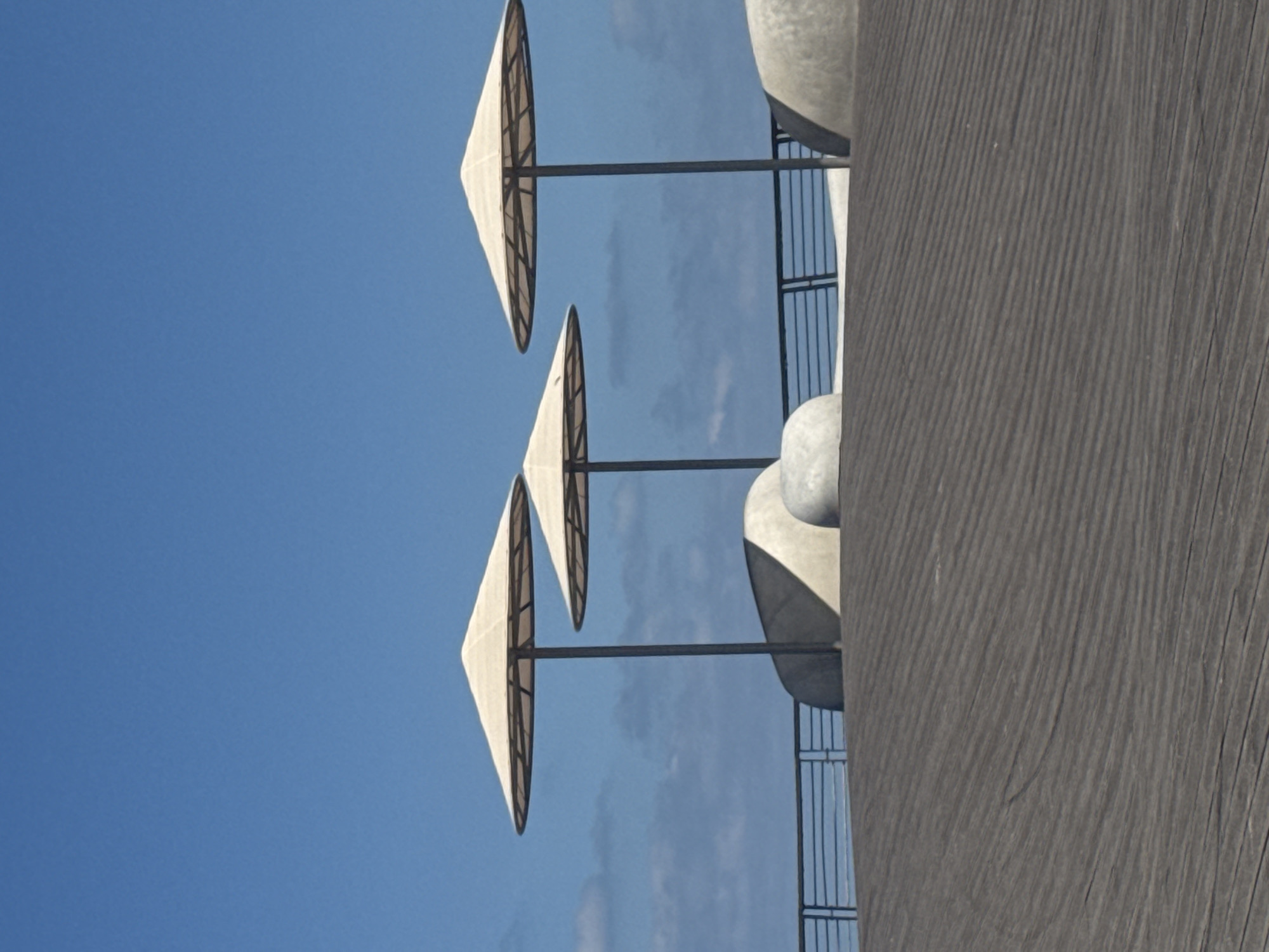 The image shows a partial view of the Dizingoff Square fountain in Tel Aviv, Israel, featuring mushroom-shaped shades atop metal poles against a clear blue sky. A textured concrete structure is visible to the right, and metal railing segments are interspersed between the mushroom shades. The image is taken from a low angle, emphasizing the upward gaze toward the sky.