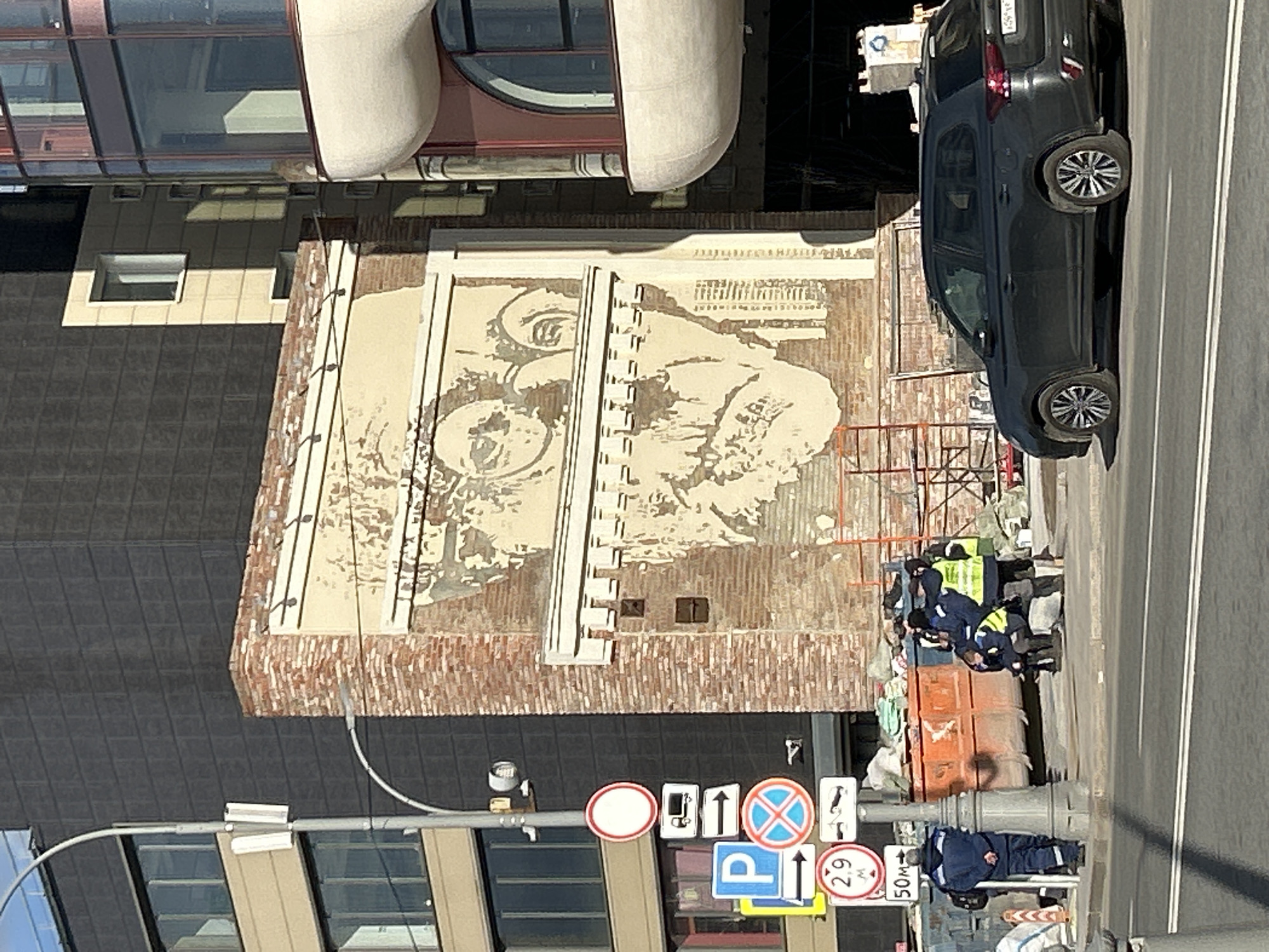 The image shows a street scene in Moscow, Russia. A large mural depicting a person with glasses is visible on the side of a brick building. A dark-colored car is parked on the side of the road. Several workers wearing blue uniforms and yellow vests are near the building, possibly engaged in maintenance or construction. Street signs are visible, including parking and traffic regulations. The background includes modern buildings with glass facades.