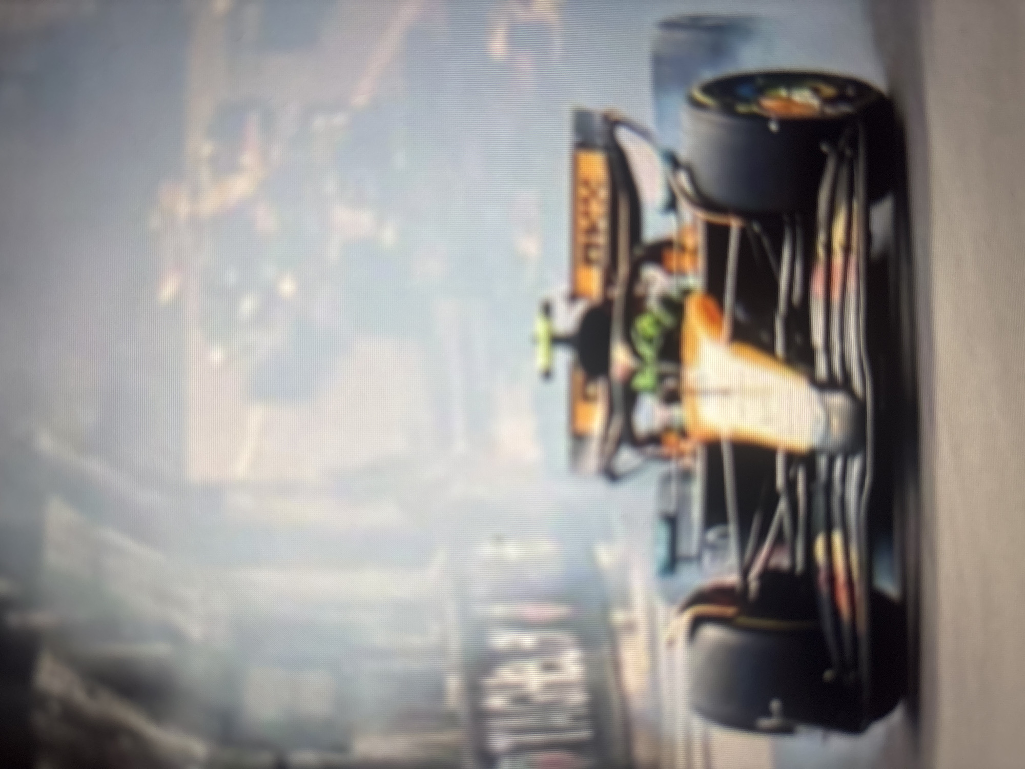 The original image, rotated 90 degrees clockwise, presents a heavily blurred and pixelated photograph of what appears to be an orange and black open-wheel racing car, likely a Formula 1 vehicle. The car is the primary subject, dominating the right side of the frame. It features large, dark-colored slick tires and discernible orange bodywork with some lighter, possibly green, accents near what would be the front of the car.
The background is extremely out of focus, showing only indistinct shapes and colors, suggesting a complex environment such as a city skyline, grandstands, or track infrastructure. This heavily blurred background, combined with the general motion blur effect, implies the car is either in motion or captured during a dynamic event like a race or practice session.
Notable details include the prominent slick tires, characteristic of racing, and the distinctive orange livery often associated with certain racing teams. The image quality is poor, with a visible pixel grid, suggesting it might be a photo taken of a digital screen. Due to the severe blur and pixelation, no specific text, logos, or identifying marks are legible on the car or in the background. The time of day and weather conditions are impossible to determine from the image.