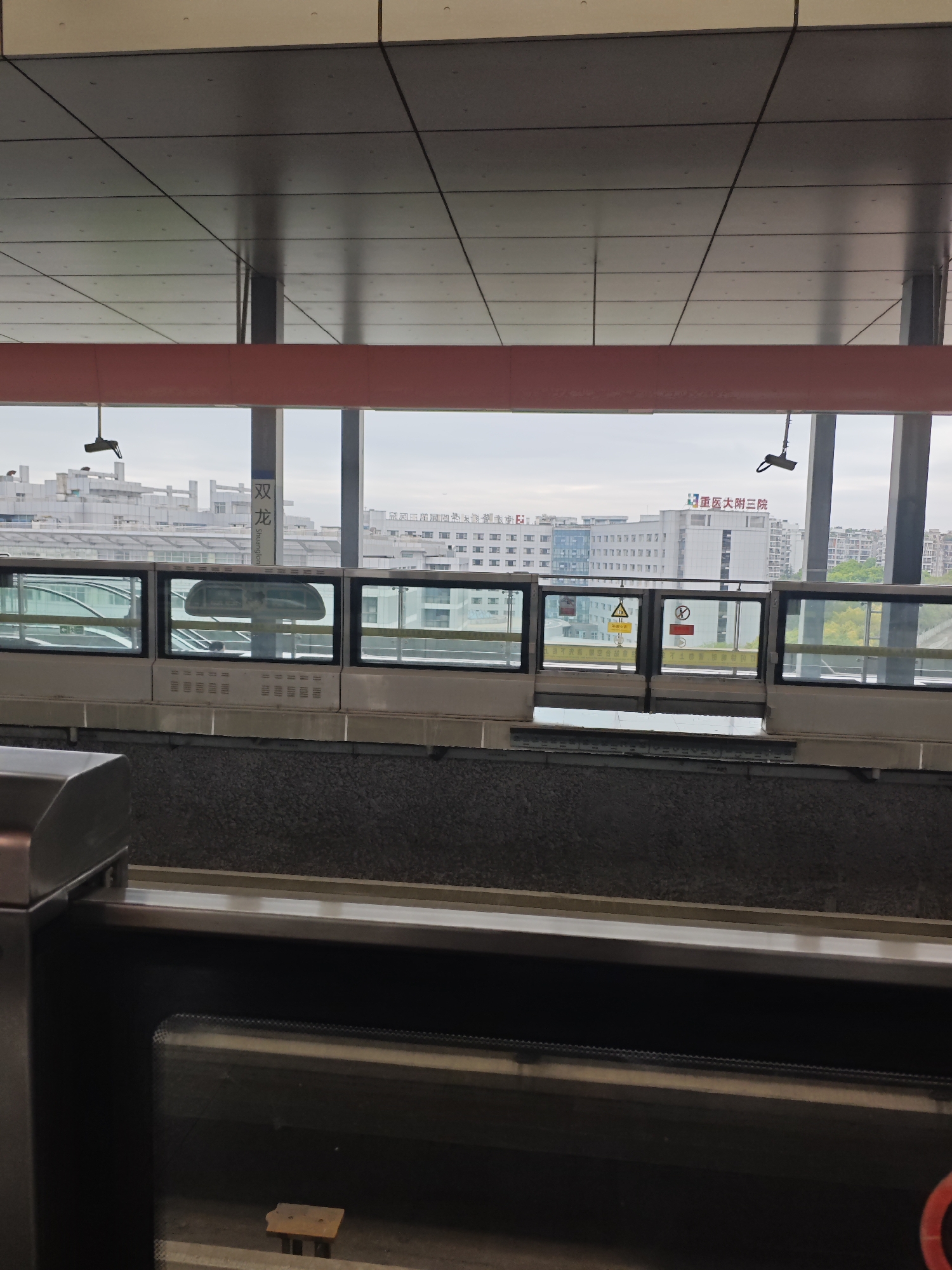 The image shows the inside of a subway station in Liangjiang, China, at the Shuanglong station. The station platform features a partially transparent barrier wall with automated doors. Through the barrier doors, one can see buildings in the cityscape, including the 重医大附三院 (Chongqing Medical University Third Affiliated Hospital). There is a red horizontal beam near the top of the frame, under a grid-patterned ceiling. A dark grey panel is near the bottom, and a partially visible structure with stainless steel and black surfaces is on the lower left. A security camera is hanging from the ceiling.