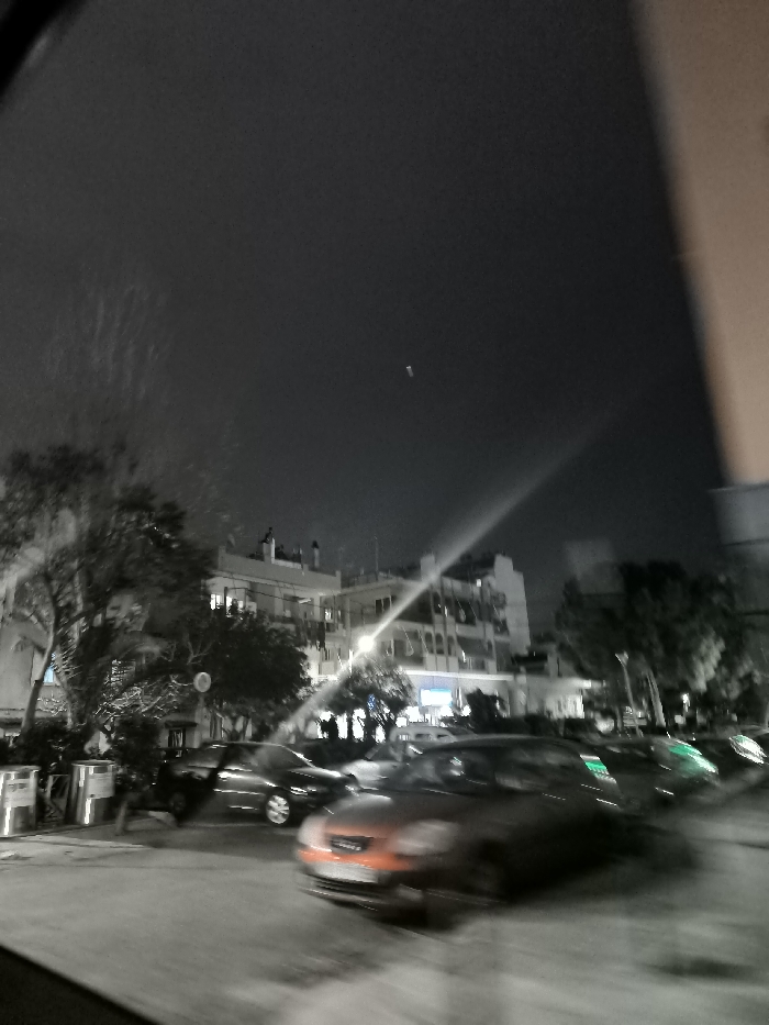 This nighttime photograph captures a street scene in Drapetsona, Greece, characterized by motion blur and a selective color effect. The sky is dark, indicating late hours, with a small, elongated, light-colored object discernible high above in the center-right, possibly an aircraft or distant light. Faint, textured streaks appear in the upper left of the sky, potentially reflecting rain or marks on a window pane through which the photo was taken.
The street below is lined with multi-story buildings, some showing illuminated windows, suggesting residential or commercial activity. Trees with bare branches are visible on the left and throughout the midground. Several cars populate the street; some appear parked, while others are heavily blurred, indicating movement, either of the vehicles themselves or the camera during exposure. A prominent orange or red car with blurred headlamps is visible in the mid-right foreground, standing out due to its color in the otherwise largely desaturated scene. A strong light flare or streak diagonally traverses the mid-ground, originating from the right side, likely from a bright streetlight or vehicle headlights. Scattered green lights are faintly visible on the right side of the street. The overall impression is a dynamic, urban nightscape. No legible text is visible in the image.