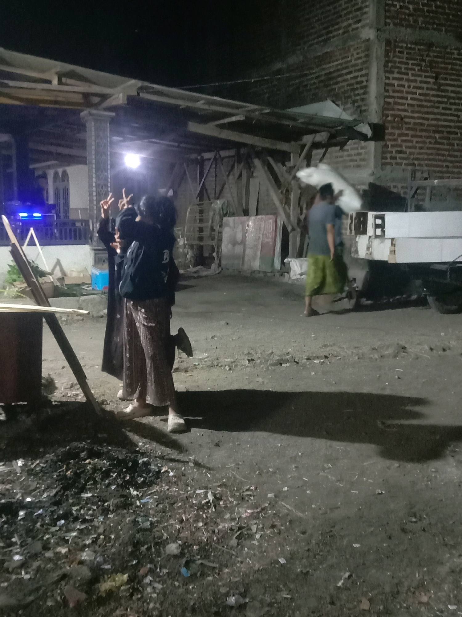 The image depicts a dimly lit outdoor scene at night in what is identified as the city of Karang Anyar, Indonesia. Two individuals are standing on the left side of the frame, with their backs partially to the camera. The person in the foreground, on the left, is wearing a dark hooded jacket and patterned pants, with their hands raised in a gesture. The second person, standing slightly behind and to the right, is wearing a dark long dress or robe. A man is visible on the right side of the frame, walking away from the camera while carrying a large white object, possibly a rolled-up tarp or fabric. He is wearing a dark t-shirt and green pants. The environment appears to be an open, unpaved area with scattered debris on the ground. Structures, including a brick wall and a wooden roof or shelter, are visible in the background. Artificial lighting, including blue lights from what appears to be a vehicle or equipment, is present. The overall impression is of a casual, perhaps work-related or social gathering, taking place outdoors at night.