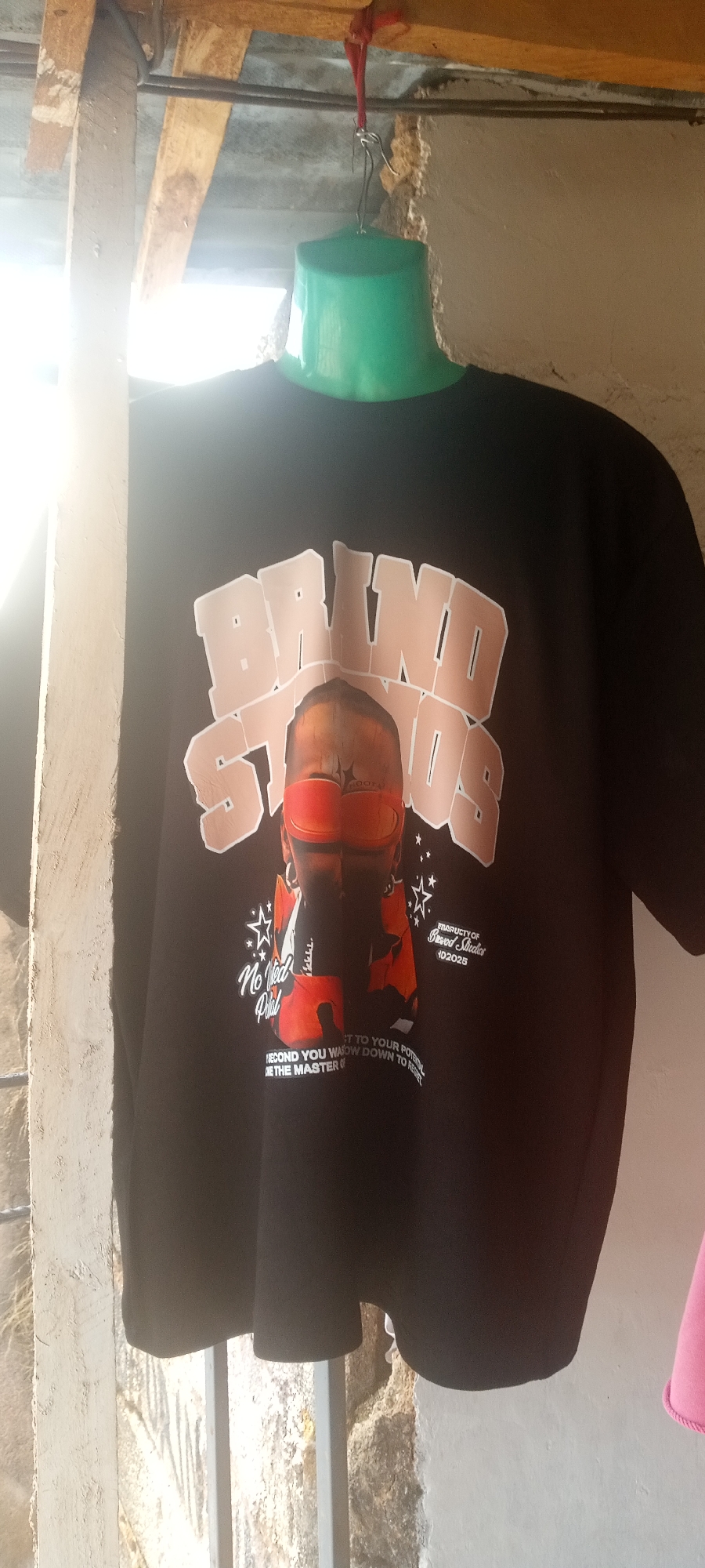 A black t-shirt with a design that says "BRUND STUDIOS" hangs from a green plastic hanger suspended by a red string. The t-shirt also features an image of a figure in red gear and lettering that includes "No Ned Phil" and "MARUCTY OF Buswod Sicclior 102025" and "SECOND YOU WASOW DOWN TO DE THE MASTER O." The t-shirt is displayed against a backdrop of weathered, light-colored walls and metal bars. The setting is outdoors, evidenced by natural light. The specific location is Jos, Nigeria.