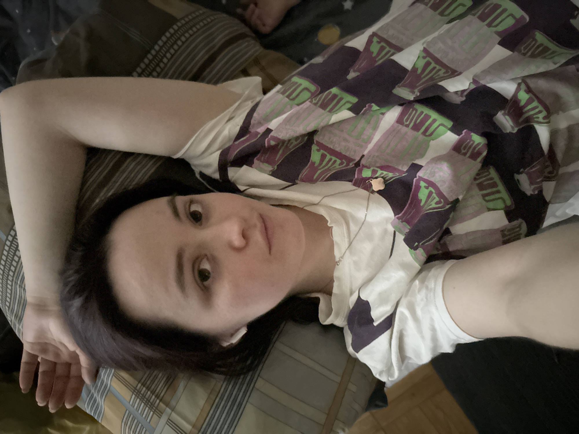 A woman with dark hair is lying on a patterned bed. She is wearing a white t-shirt with a design of what appears to be soda bottles in shades of purple and green. Her left arm is extended behind her head. The location is indeterminate and cannot be verified as Izhevsk, Russia.