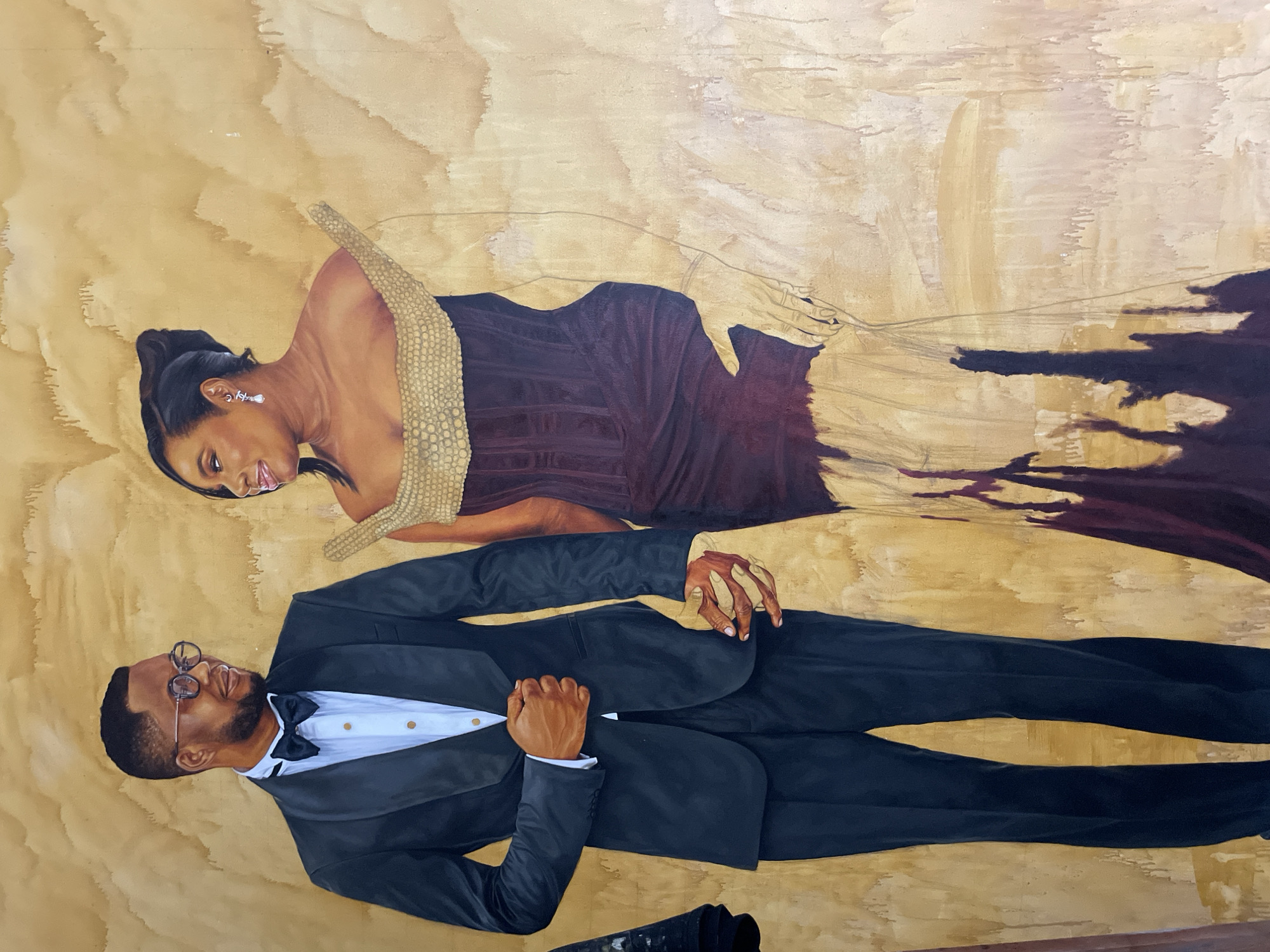 This is a full-length oil painting of a Black man and woman, likely a couple, standing side-by-side. The background is a textured, abstract wash of beige and brown tones, reminiscent of aged parchment or canvas.
The man, on the left, is wearing a black tuxedo with a black bow tie and a crisp white shirt. He has short, dark hair, a beard, and is wearing glasses with dark rims. His left hand is tucked inside the lapel of his jacket, and his right hand is held by the woman's. He is looking to his right, towards the woman, with a slight smile.
The woman, on the right, is wearing a striking, off-the-shoulder gown. The bodice appears to be a dark plum color, and it's adorned with a textured, gold, almost scale-like embellishment around the neckline. The lower part of the dress is unfinished, with the plum color giving way to lighter tones and appearing to be in a state of partial rendering or artistic interpretation. She has her hair styled up, and she is wearing dangling earrings. She is looking towards the man, with her tongue slightly protruding, suggesting a playful or intimate moment. Her left hand is holding the man's right hand.
The painting is detailed, with attention paid to the textures of the clothing and the skin tones of the subjects. The style is realistic, with a touch of artistic flair in the rendering of the woman's dress and the background. There are no specific architectural elements or landmarks visible in the painting that would indicate a precise location within Ado, Nigeria. The scene is intimate and focused on the couple.