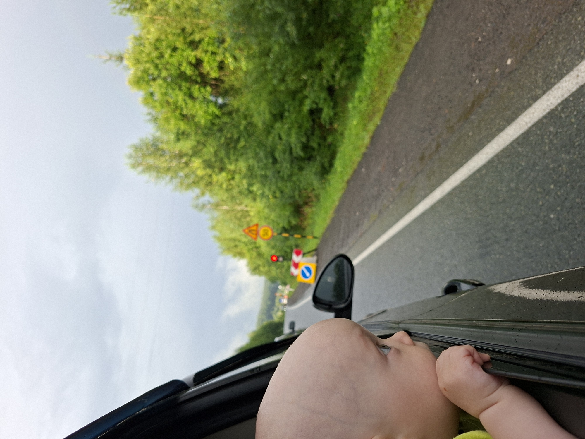 A young child, with light skin and sparse light hair, leans out of a car window, looking towards a rural road in Yugo-Kamskiy, Russia. The child's head and shoulder are visible in the foreground, resting on the car door frame. The car is traveling along a two-lane asphalt road that curves to the left. The road is lined with lush green trees and vegetation on both sides. In the distance, there are several road signs, including a yellow triangular sign with a red border and a black symbol of falling rocks, a circular sign with the number 50 in a red circle, and traffic lights. There are also construction barriers, including a yellow and blue sign indicating a detour or lane closure. The sky is overcast with gray clouds, and the road surface appears wet, suggesting recent rain. The overall scene captures a moment of travel through a scenic, somewhat inclement, rural landscape.