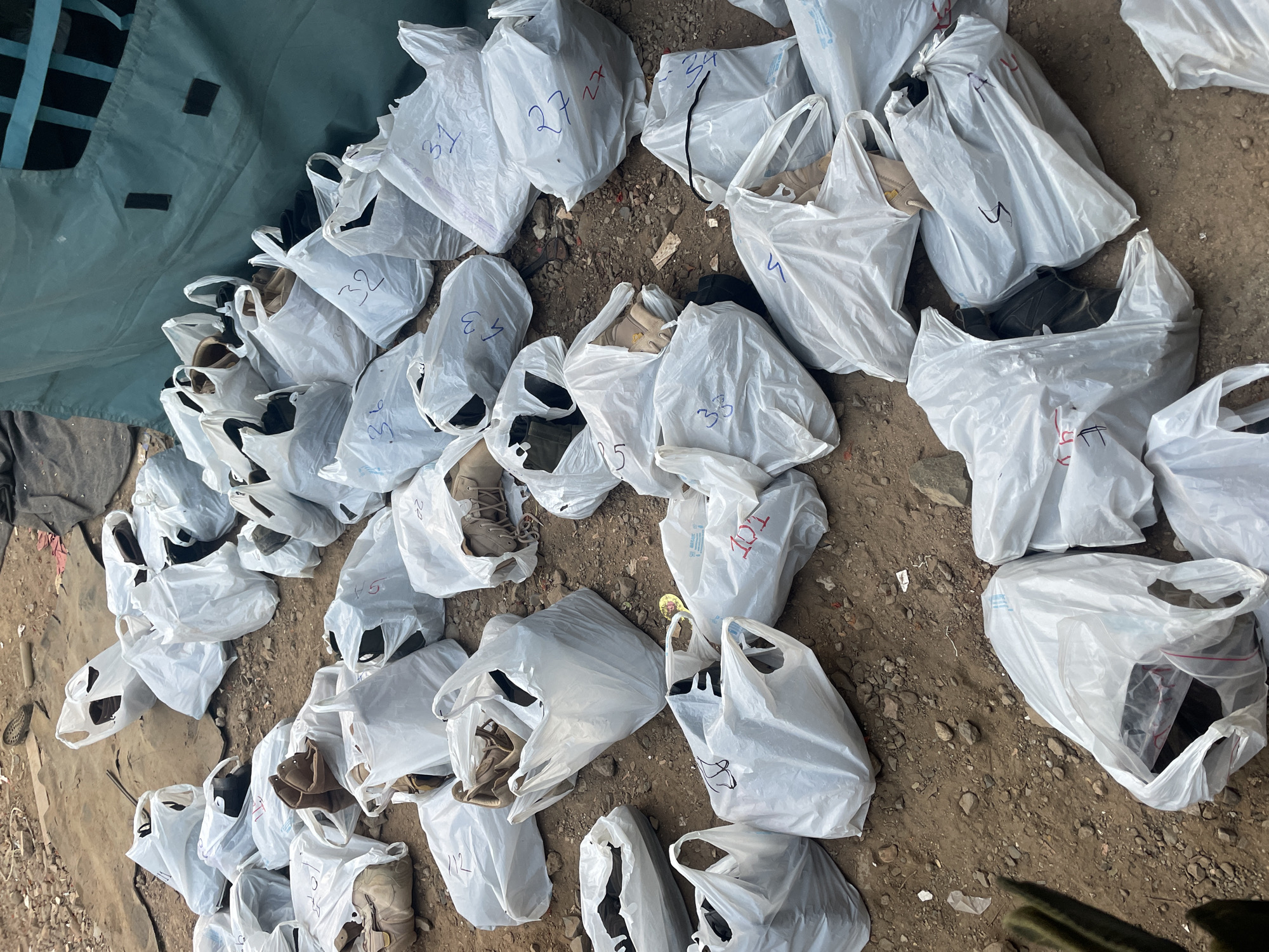 The image shows numerous white plastic bags scattered on a dirt and rock-strewn ground. Many of the bags are marked with numbers. Most bags appear to contain shoes, as glimpses of different shoe styles and colors are visible within the bags. The ground is uneven and appears to be outdoors. A section of green fabric, possibly part of a tent or tarp, is visible in the upper left corner. The location, Mira-Bhayander, India, is not discernible from the image.