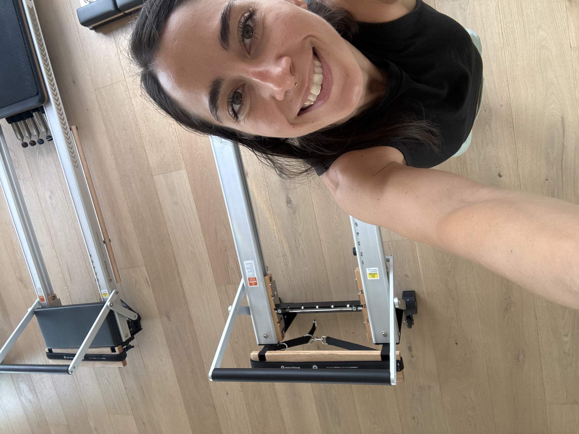 In this vertical, wide shot, a smiling woman is taking a selfie in what appears to be a Pilates studio. The floor is a light-colored wood, and two Pilates reformers are visible. The woman is wearing a black t-shirt and has dark hair. Her face is close to the camera, and she is looking directly at it with a wide smile, revealing white teeth and a gold nose ring. Her left arm is extended to capture the photo.
The Pilates reformers are silver with black padded seats and foot bars. One reformer in the foreground has a label on its side that reads "MERRITHEW." Another label, partially visible, says "PILATES REFORMER EQUIPMENT." The reformers are positioned on the wooden floor.
The image has a bright, natural light, suggesting it was taken during the daytime. The setting seems to be indoors, possibly a home studio or a commercial Pilates facility. The woman's expression conveys happiness and confidence. The image's orientation is portrait, with the top of the frame showing the woman's forehead and hair, and the bottom showing the equipment.
