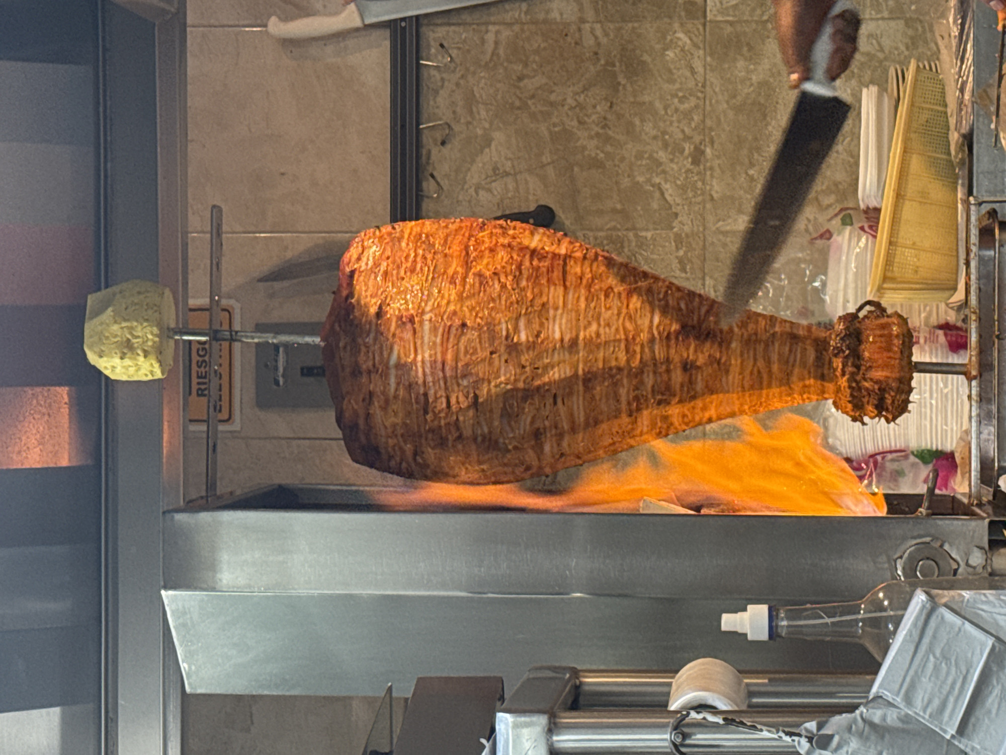 In Puebla, Mexico, a vertical shawarma-style meat roasting machine cooks meat over a flame. A chef or worker in the upper right wields a large knife, ready to shave cooked meat from the rotating spit. The meat is layered and conical in shape. A large piece of pineapple is affixed to the spit on the far left. Below the spit, a metal basin catches drippings and likely charred bits. Below that, plastic-wrapped items are visible through the metal frame.