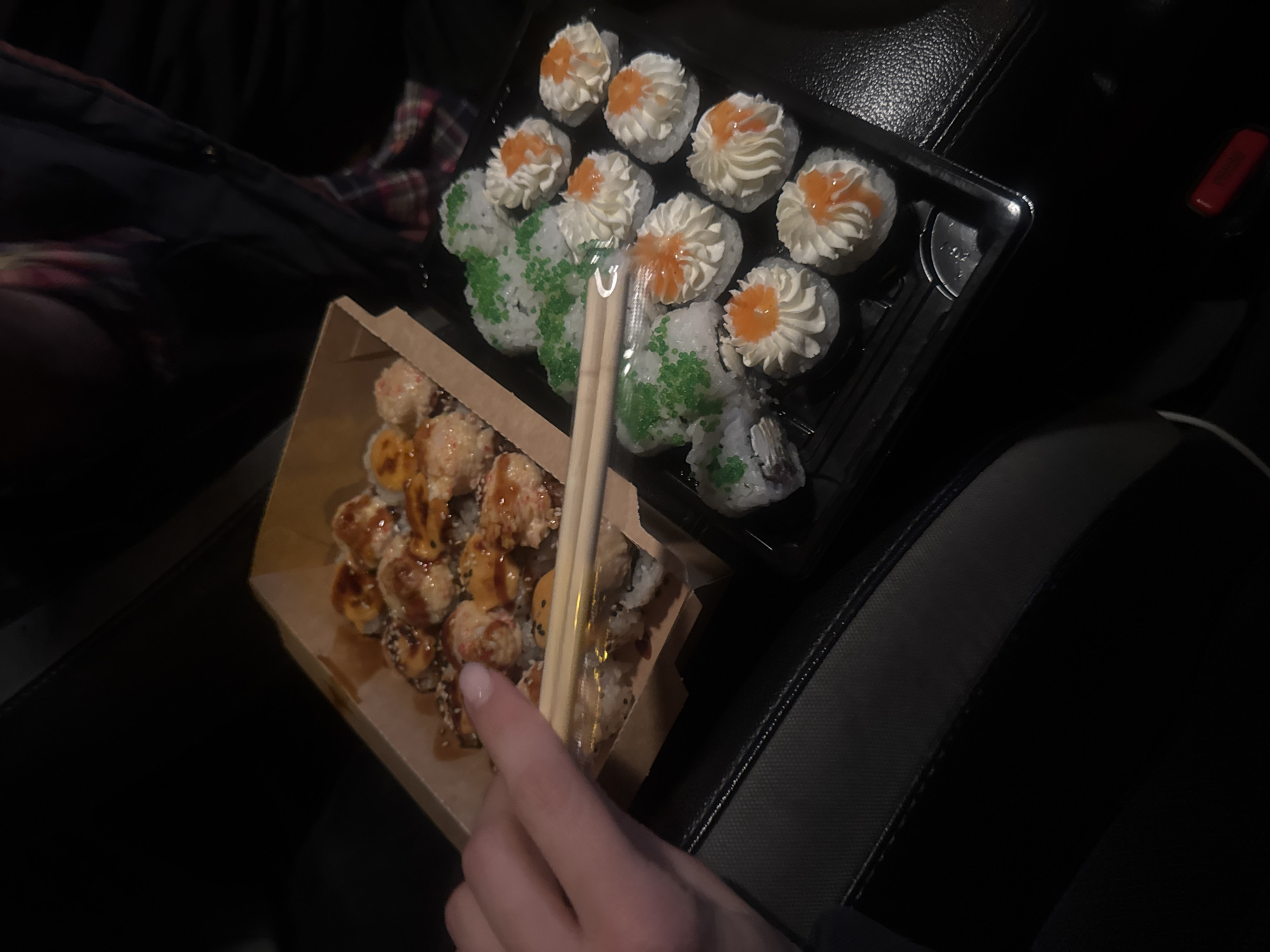 The image shows two containers of sushi on a dark surface, likely a car seat. A person's hand is visible, holding chopsticks near a carton of sushi. The sushi in the carton appears to be baked rolls with a sauce and sesame seeds. The other container, made of black plastic, contains sushi with green tobiko and another type topped with cream and orange sauce. The location of the image is difficult to determine from the image itself, and there is no information to suggest it is in Tbilisi, Georgia. The only clue is the presence of sushi in the image.