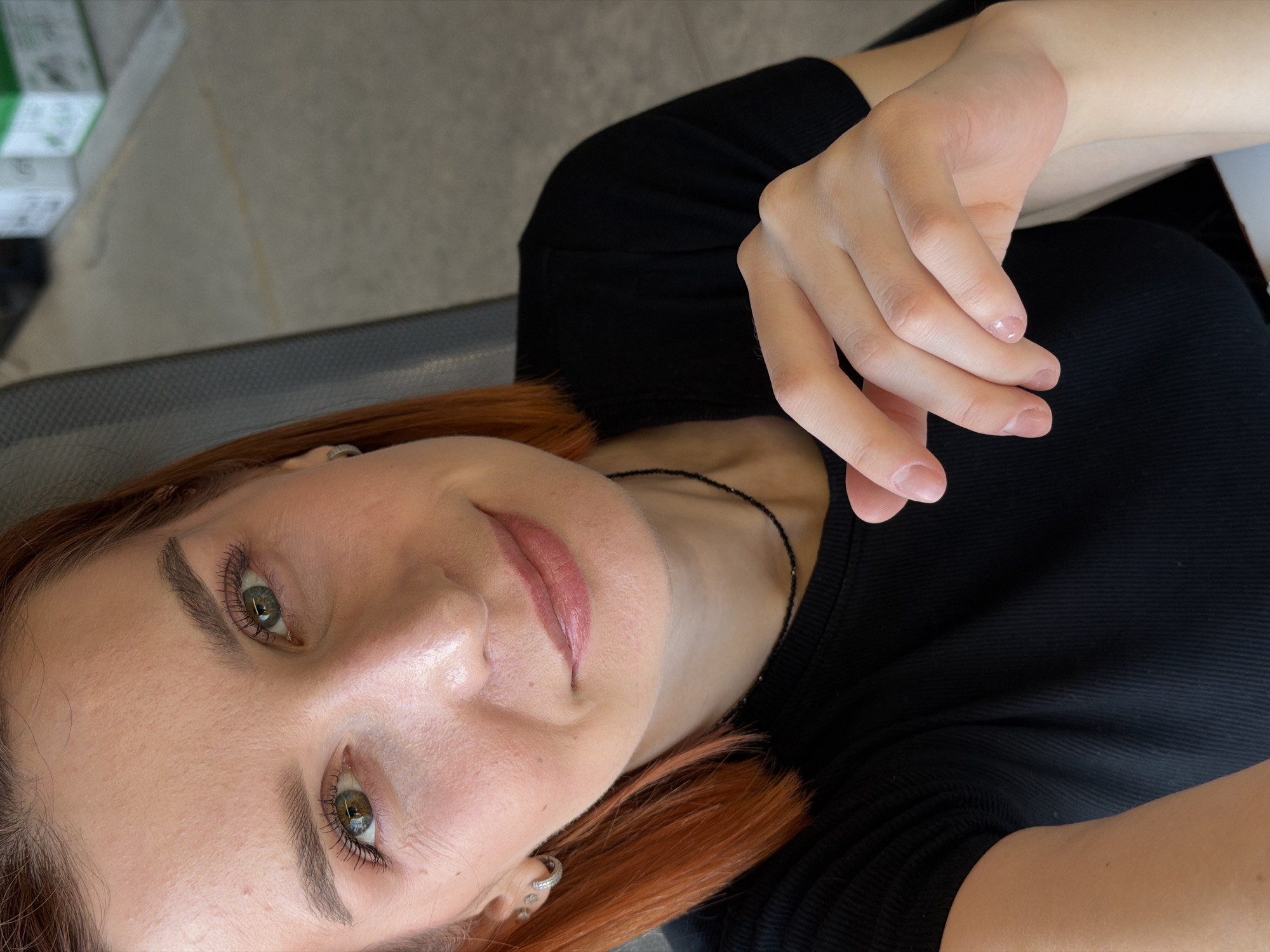 The image depicts a woman with red hair and fair skin. She has green eyes and is wearing a black top. Her hand is raised near her face. The background is blurry and appears to be an interior space. There is a metallic surface visible in the background, and possibly boxes. The location is specified to be Chaikovsky, Russia.