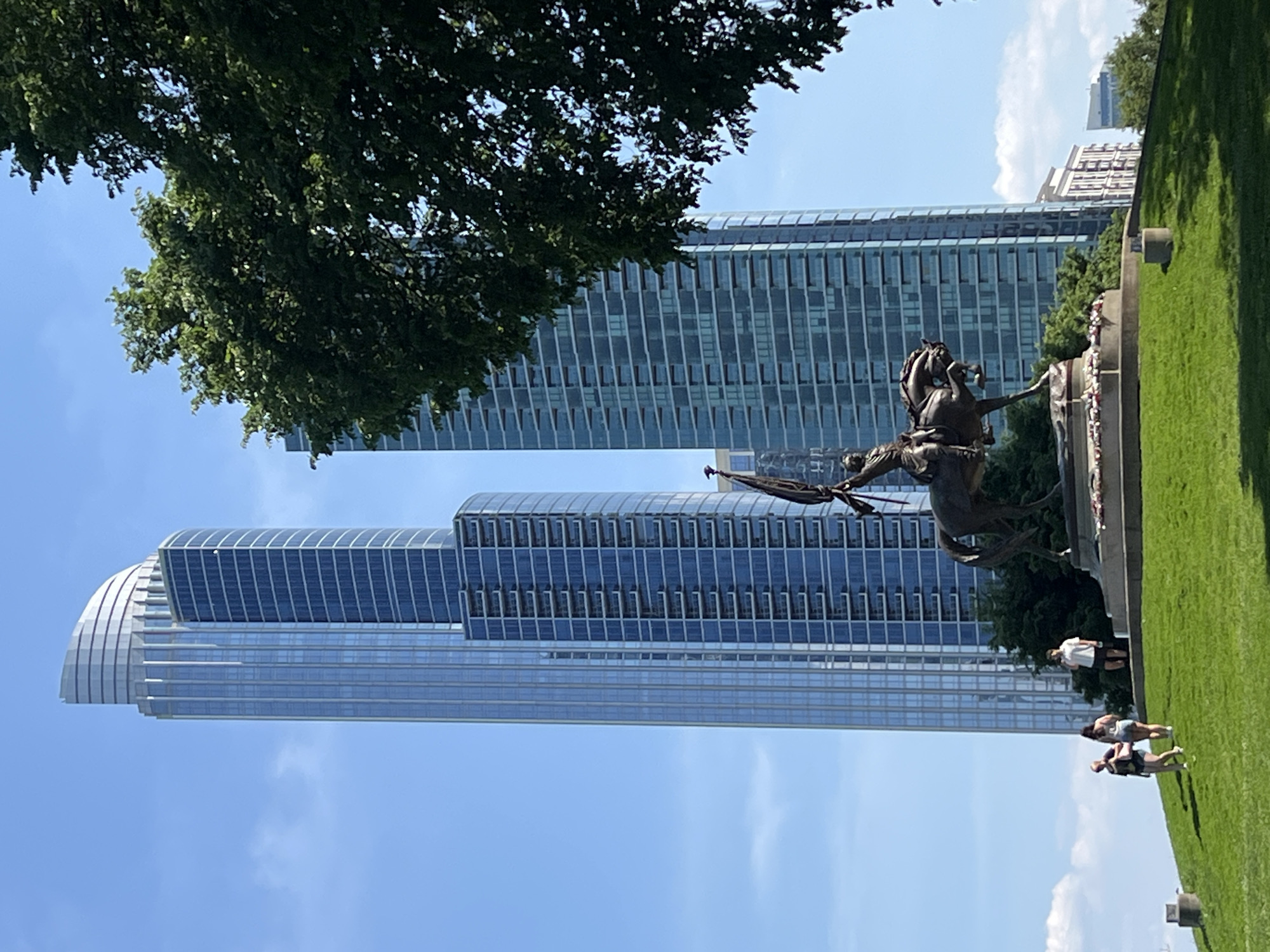 The image depicts a statue of a person on horseback, located in a park with modern skyscrapers in the background. The statue is made of bronze and appears to be in a historical style. Lush green grass covers the park area, with several people standing nearby. Two modern skyscrapers with glass facades dominate the background, indicating the location is in an urban setting. The sky is light blue with some clouds. The statue and buildings are partially obscured by tree foliage on the left side of the frame. The image is taken in daylight and appears to be outdoors. The city where this is located is Unknown, Unknown.